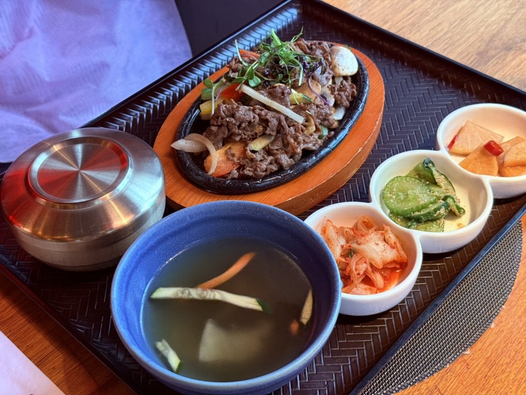 YORI Vienna bulgogi baekban set - sizzling cast-iron platter of thinly-sliced marinated beef bulgogi with mushrooms onions and carrots on a wooden trivet, beside a covered stainless rice bowl, a clear soybean-sprout soup in a blue bowl, and a 3-banchan tray with pickled daikon, sesame cucumber salad and kimchi