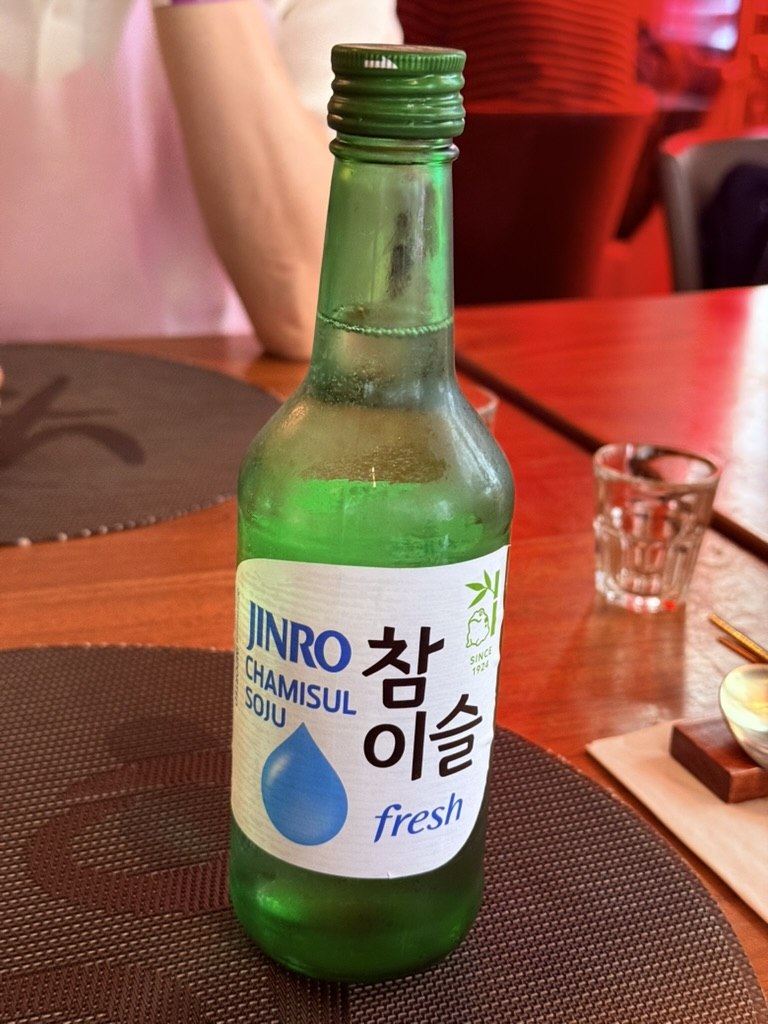 Green bottle of Jinro Chamisul fresh Korean soju with white label showing 참이슬 calligraphy and a small drop logo, on a wooden table at YORI Vienna with a small empty shot glass beside it