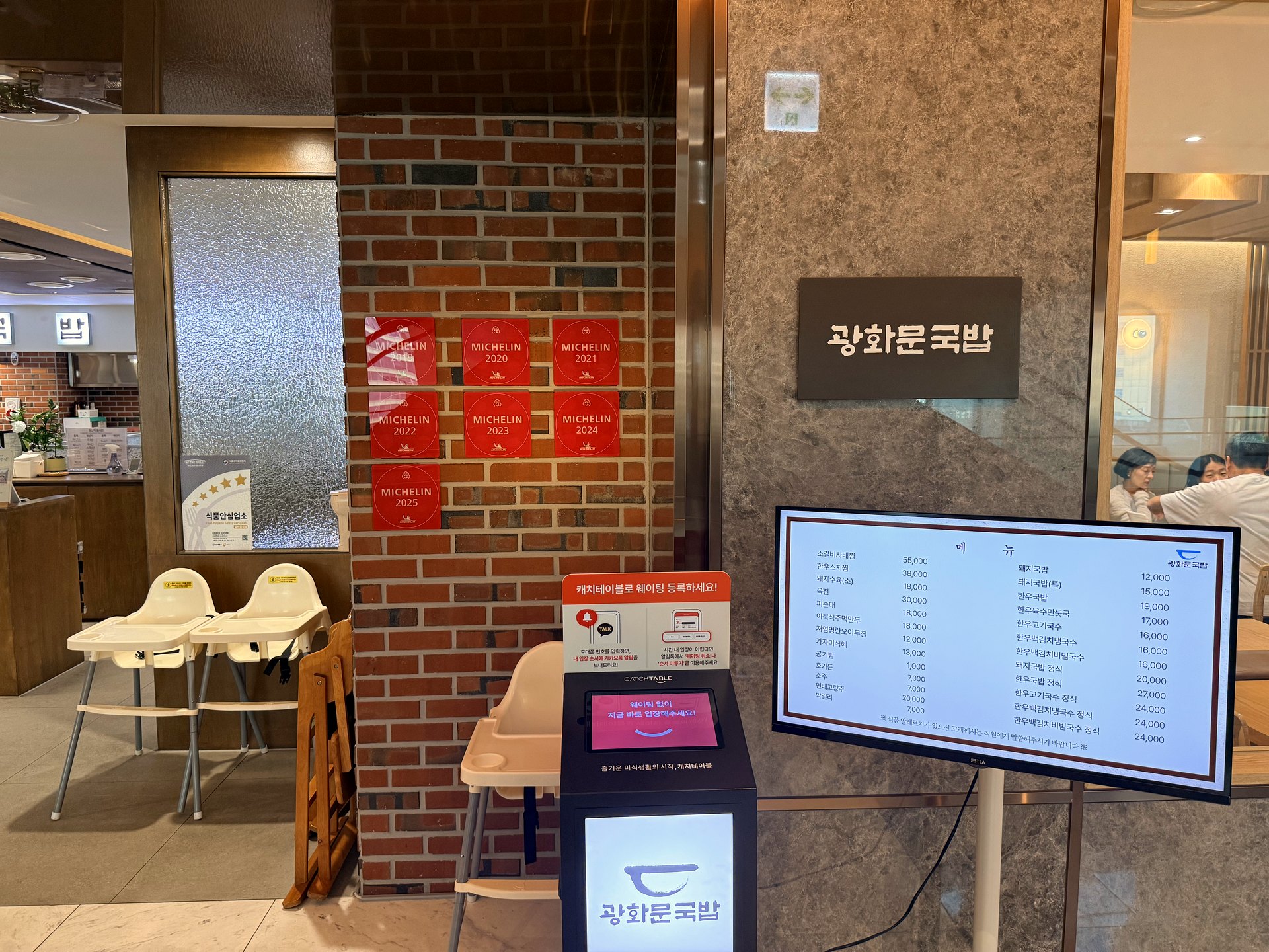 Entrance of Gwanghwamun Gukbap at Shinsegae Myeongdong - a brick pillar covered with red MICHELIN plaques for years 2019 through 2025, next to a dark grey sign reading 광화문국밥 in Korean, a CatchTable waitlist kiosk, baby high chairs, and a digital menu screen on the right