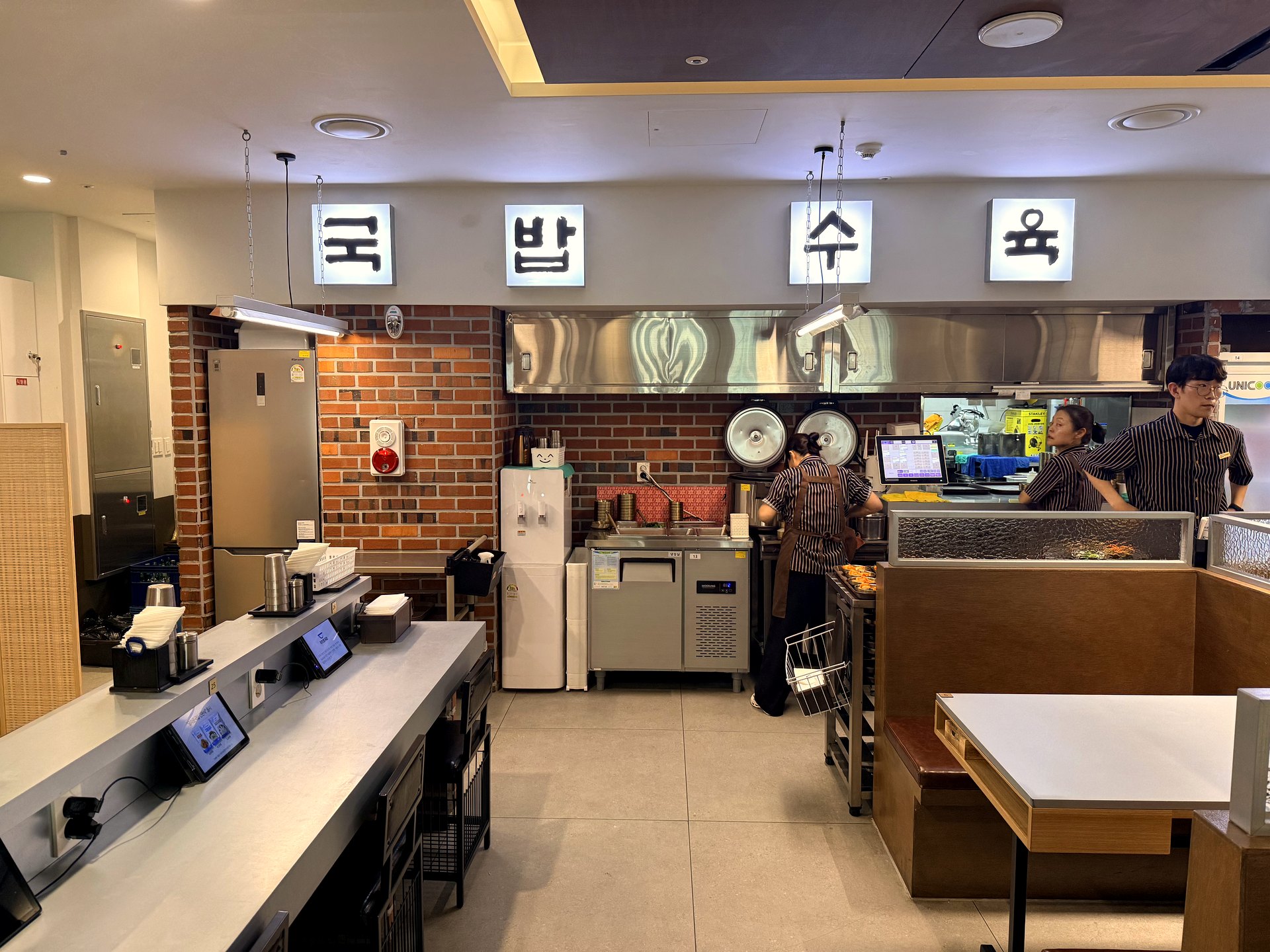 Interior of Gwanghwamun Gukbap Shinsegae Myeongdong - an open kitchen with exposed red brick walls, staff in brown striped aprons working behind a low counter, large brass broth pots, a water dispenser, and glowing white light-box Korean letters reading 국밥 국수 육 (gukbap, noodles, meat) hanging from the ceiling