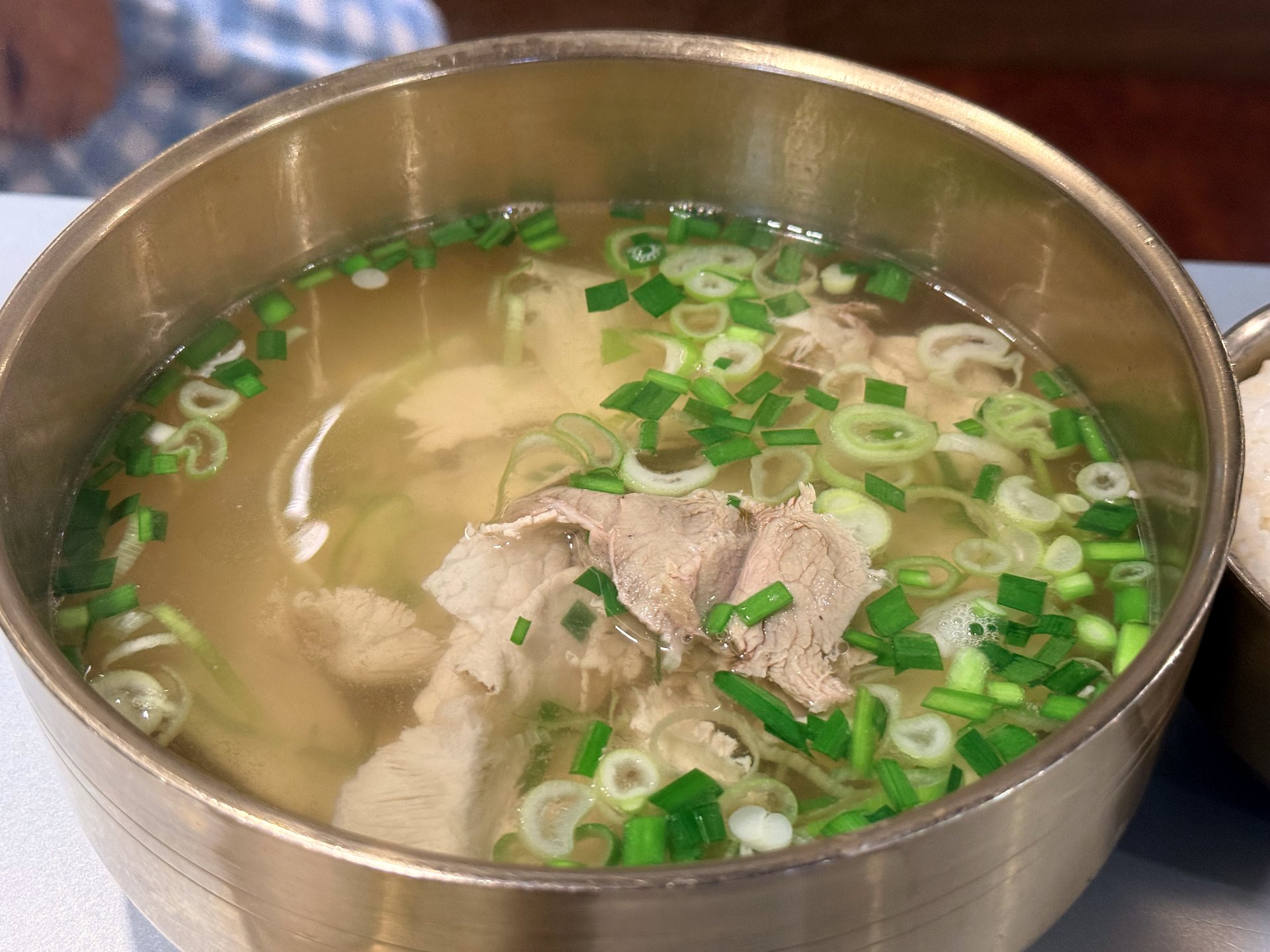 Clear pork gukbap at Gwanghwamun Gukbap Shinsegae Myeongdong - a brass bowl of light, translucent pork broth with thin slices of lean boiled pork and a heavy topping of chopped green scallions and chives, a clean and non-greasy style of Korean pork soup, with a bowl of rice beside it