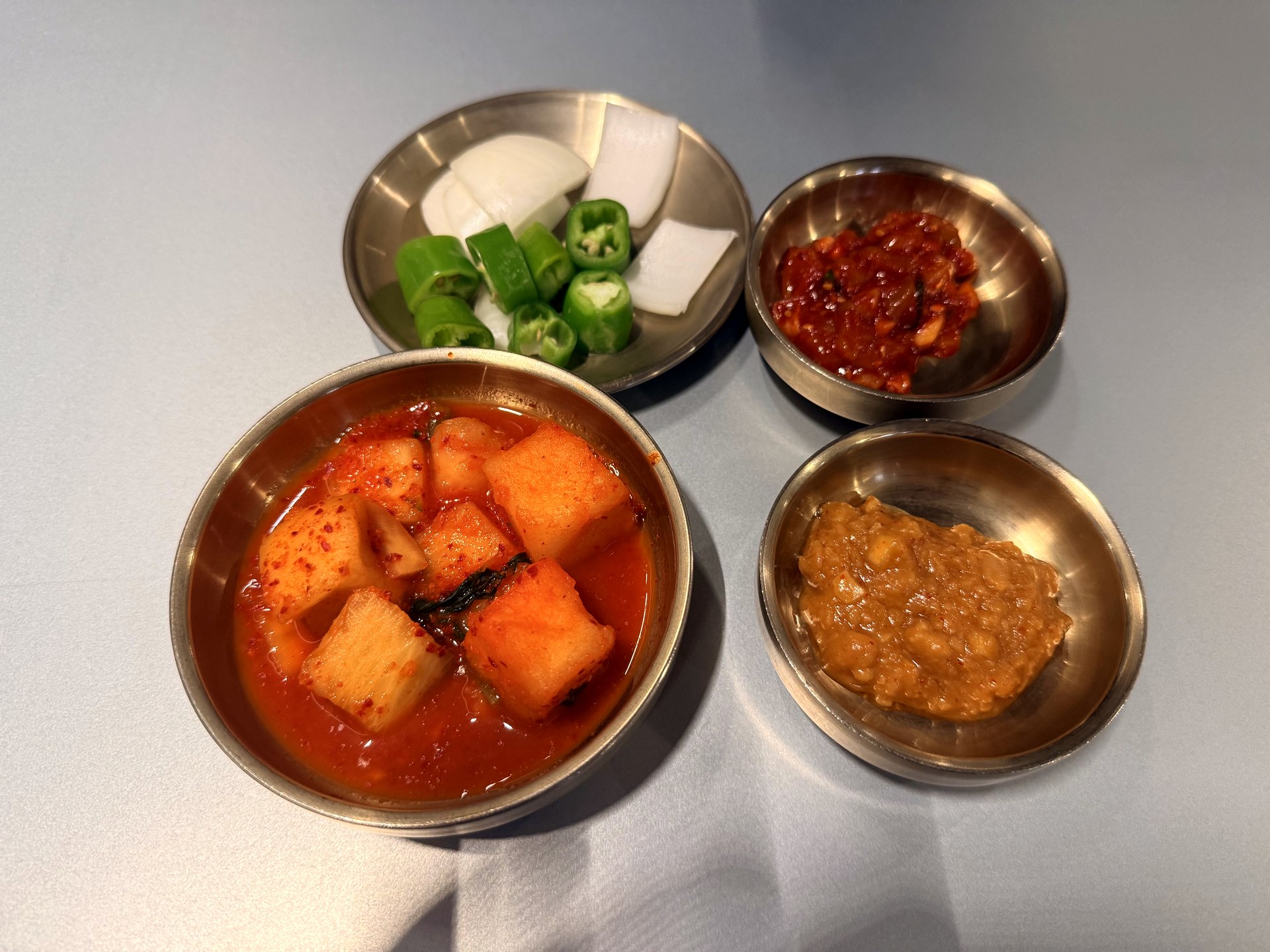Banchan at Gwanghwamun Gukbap Shinsegae Myeongdong - four small brass dishes holding cubed radish kimchi in red seasoning, sliced raw onion with cut green chili peppers, a red chili-paste ssamjang, and a brown fermented soybean side, arranged on a grey table