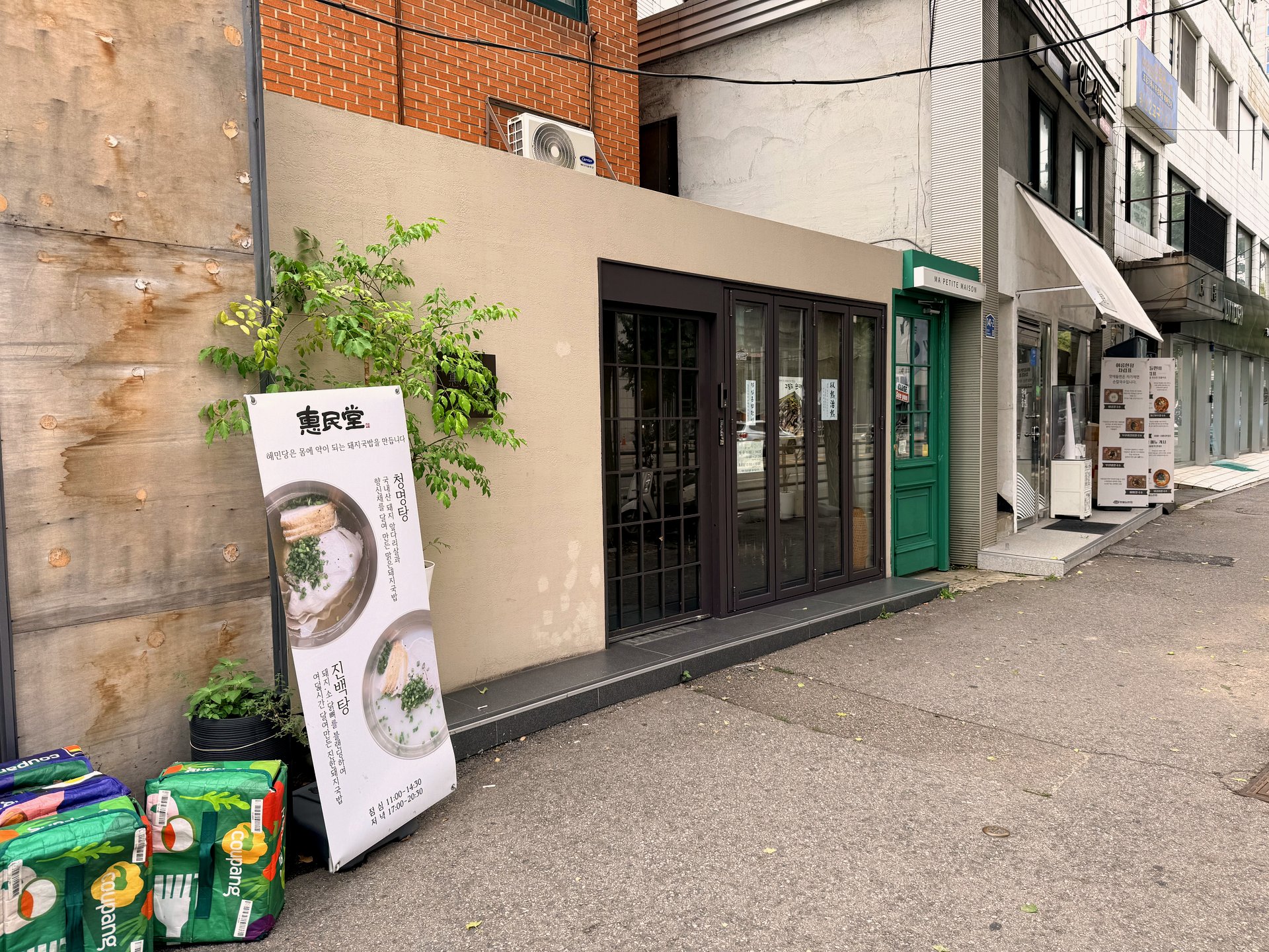 Exterior of Hyemindang in Yongsan - a beige single-story storefront with brown folding-glass doors and a small vertical banner reading 惠民堂 Hyemindang with photos of cheongmyeongtang and jinbaektang and hours, on a quiet side street with Coupang delivery bags stacked outside