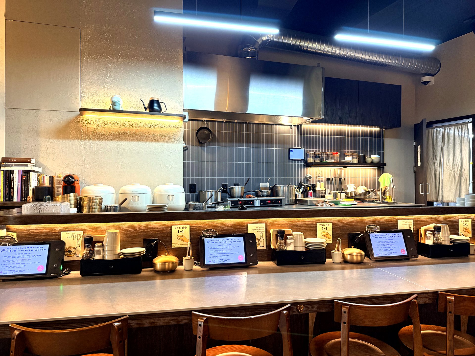 Interior of Hyemindang Yongsan - a long light-grey counter with wooden bar stools facing an open kitchen, rice cookers and brass bowls stacked on a shelf, ordering tablets along the counter, cookbooks on a side shelf, and blue LED strip lighting on the ceiling