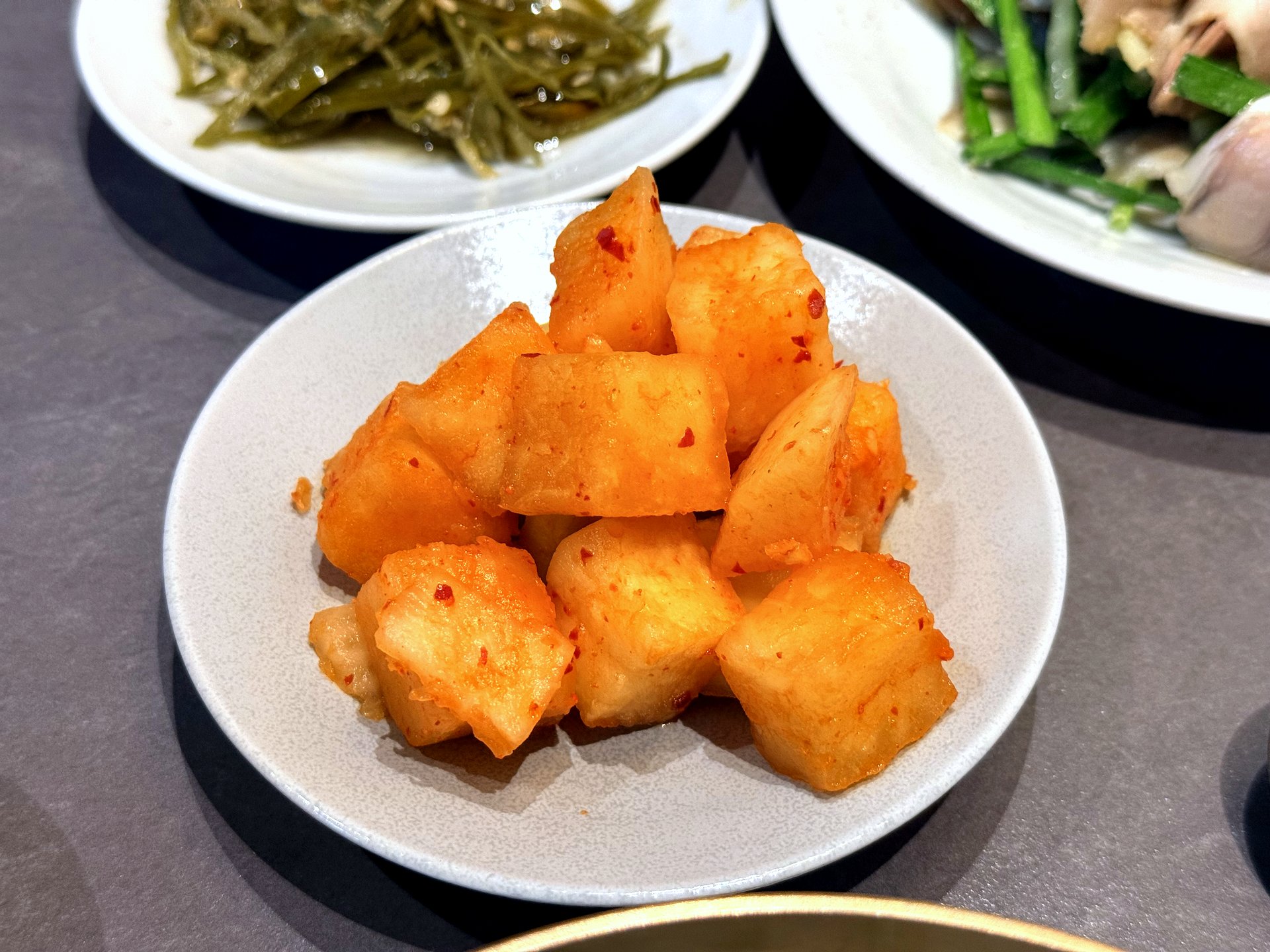Banchan at Hyemindang Yongsan - a small white bowl of glossy red-orange cubed radish kimchi (kkakdugi) in the foreground, with a plate of seasoned seaweed stems and the pork suyuk plate visible behind on a grey table
