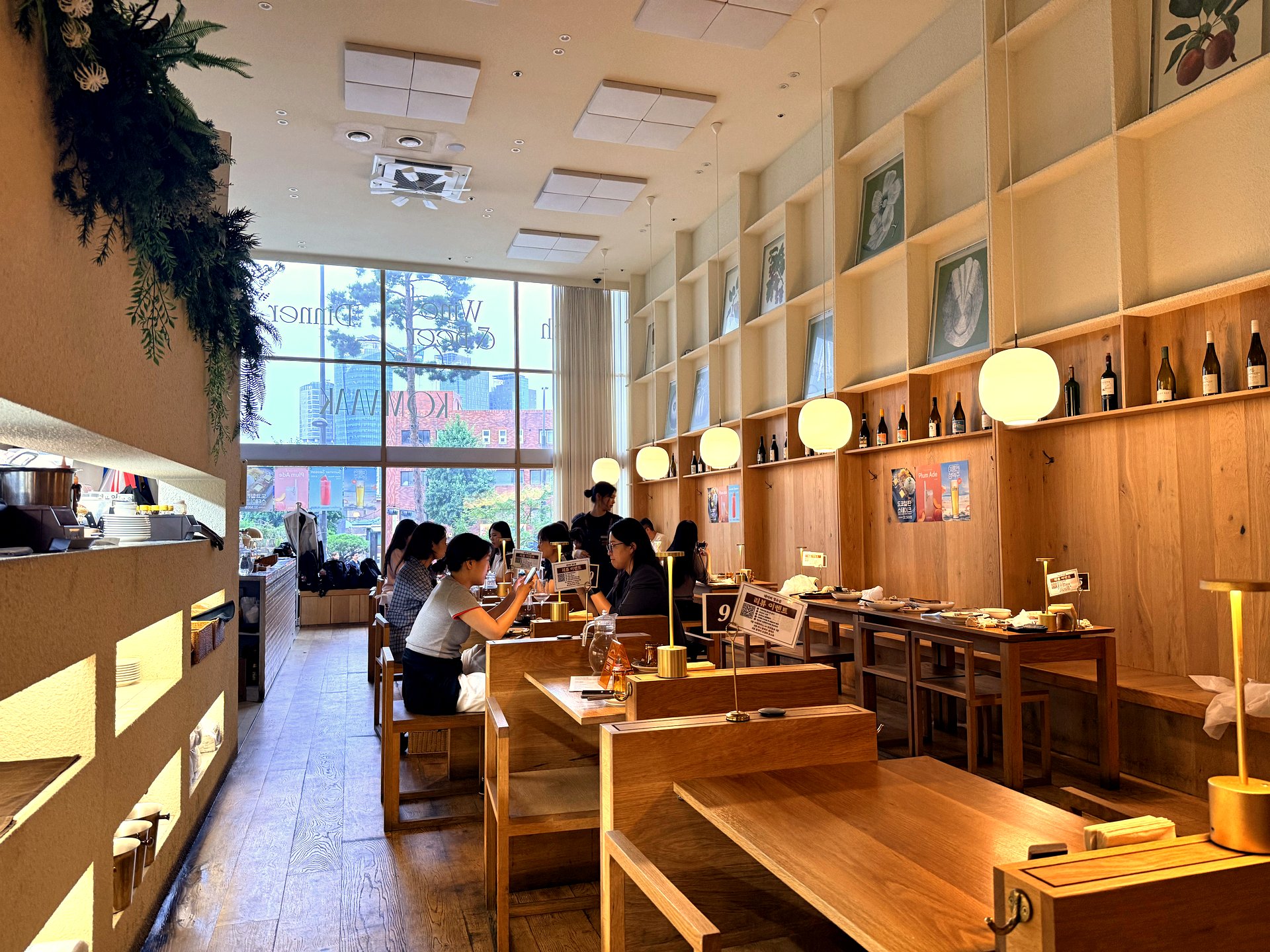 Interior of Komvaak near Seoul Station - a bright, high-ceilinged dining room with tall windows, warm wood tables and chairs, round white pendant lights, shelves lined with wine bottles and framed botanical prints, and diners seated along the window