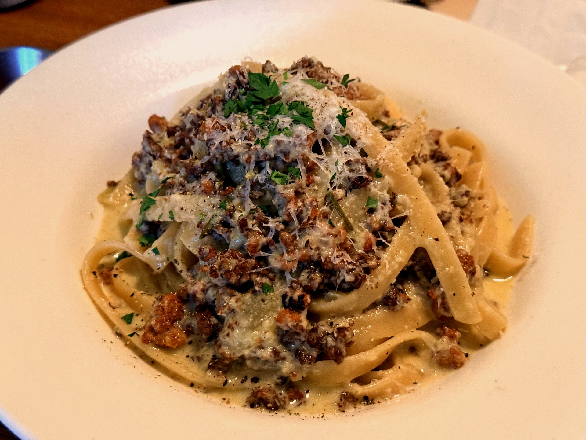 White ragu pasta at Komvaak - a wide white bowl of fresh ribbon pasta coated in a creamy pale sauce with crumbled browned sausage and beef, grated parmesan, chopped parsley and cracked black pepper