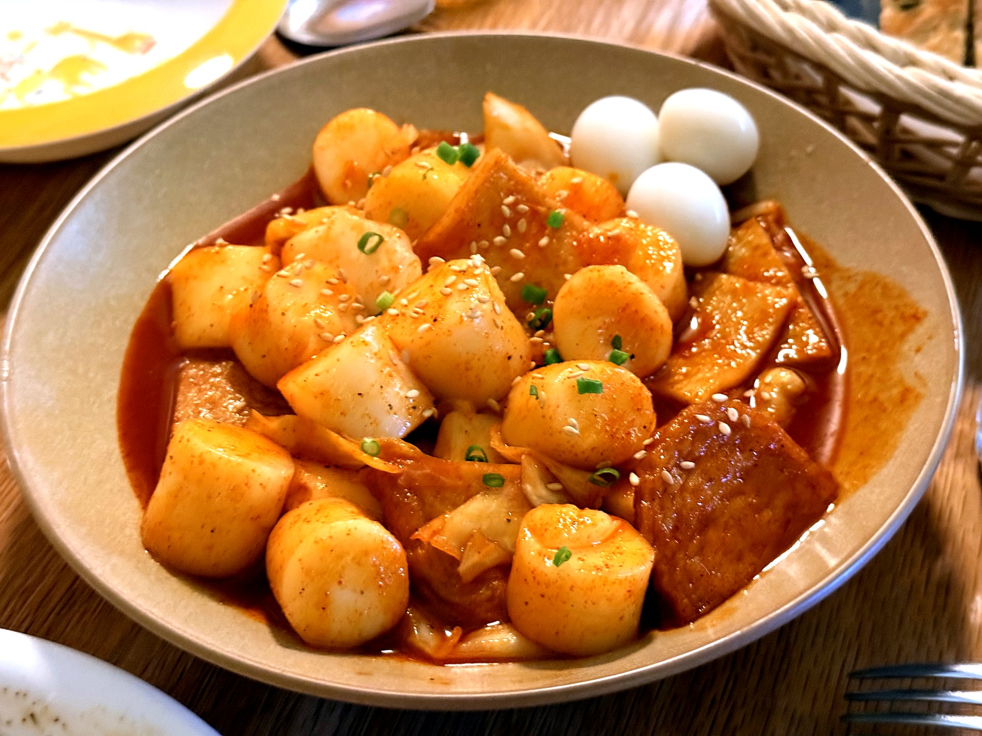 Komvaak tteokbokki - a shallow bowl of plump white cylindrical rice cakes and fish cake in a glossy red-orange sweet-and-spicy sauce, topped with sesame seeds, chopped scallion and three quail eggs