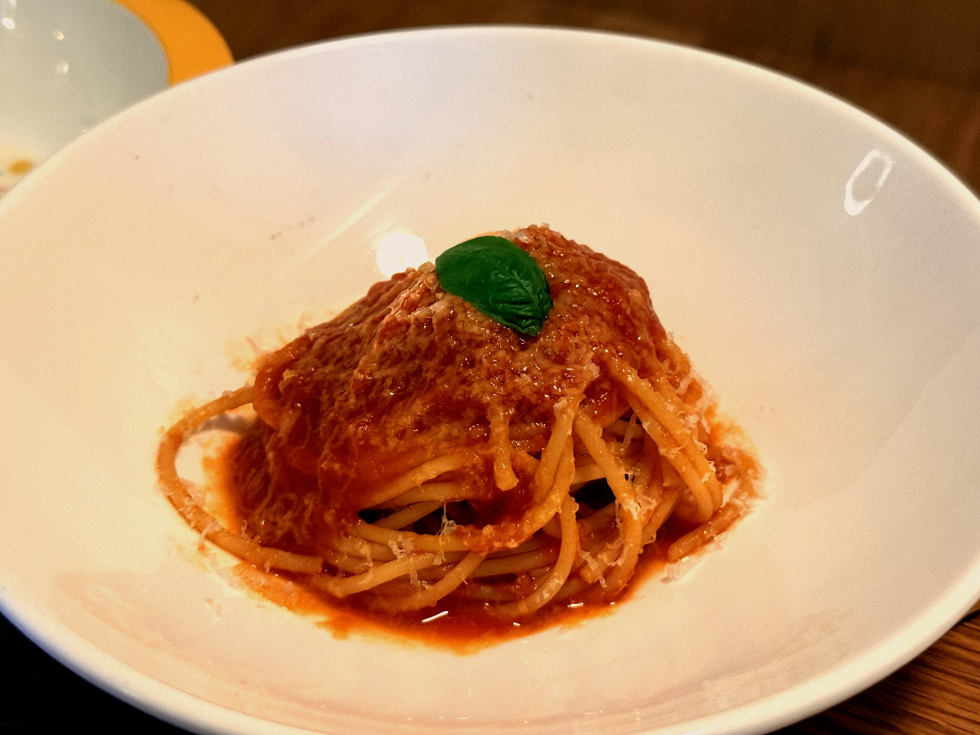 A simple tomato pasta at Komvaak - a white bowl of spaghetti in a glossy red tomato sauce with grated cheese and a single fresh basil leaf on top, the plain kid-friendly option