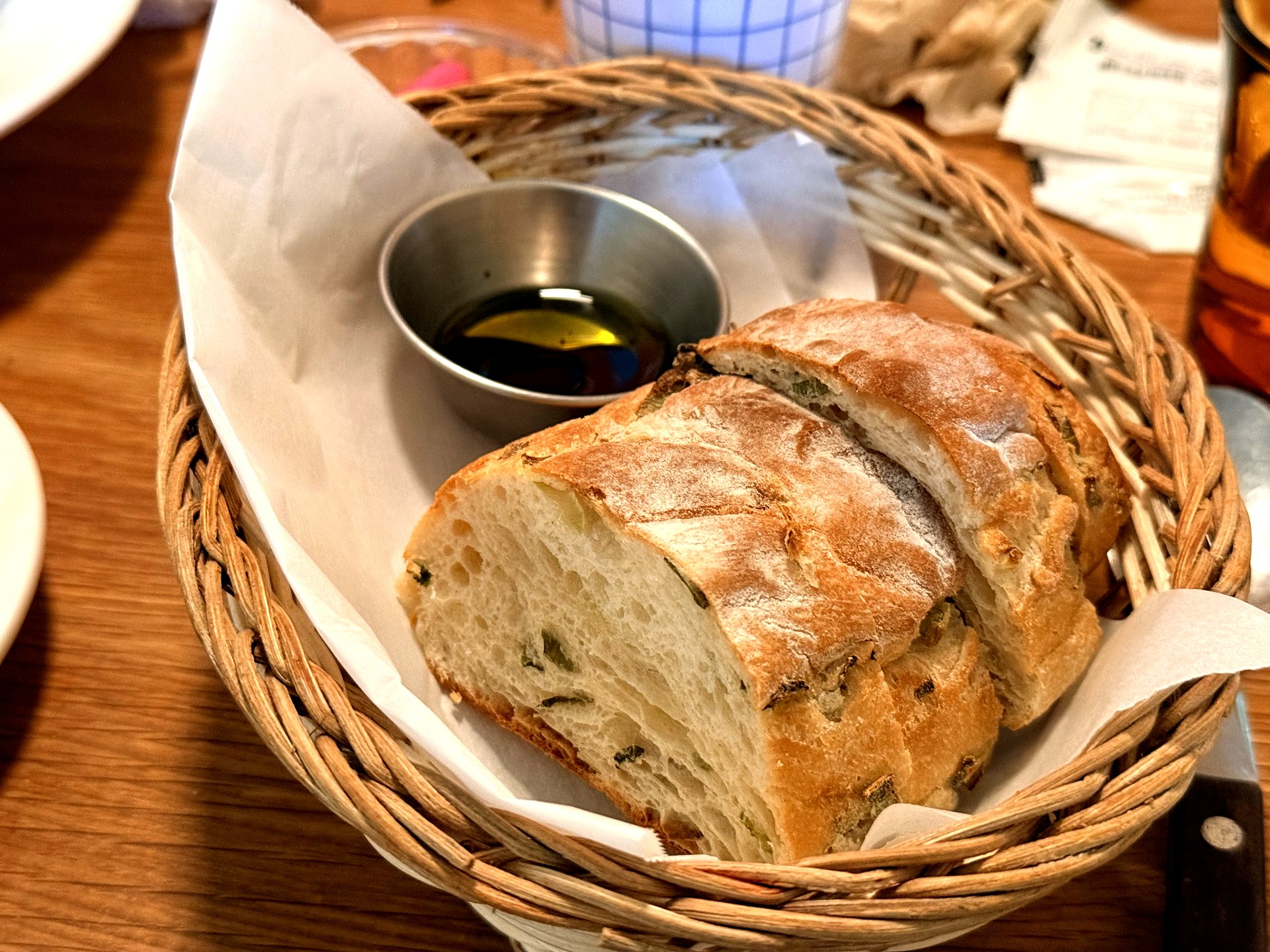 Complimentary bread basket at Komvaak - a rustic wicker basket lined with paper holding thick slices of crusty green-onion bread beside a small dish of olive oil and balsamic vinegar