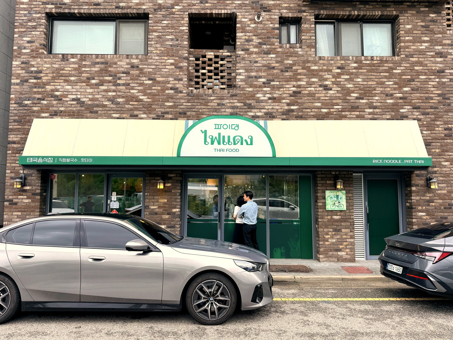 Exterior of Pai Deng in Yeongjongdo - a brown brick building with a green awning and a round sign reading 파이뎅 in Korean and ไฟแดง in Thai with THAI FOOD underneath, and side text reading Rice Noodle Pat Thai, cars parked out front
