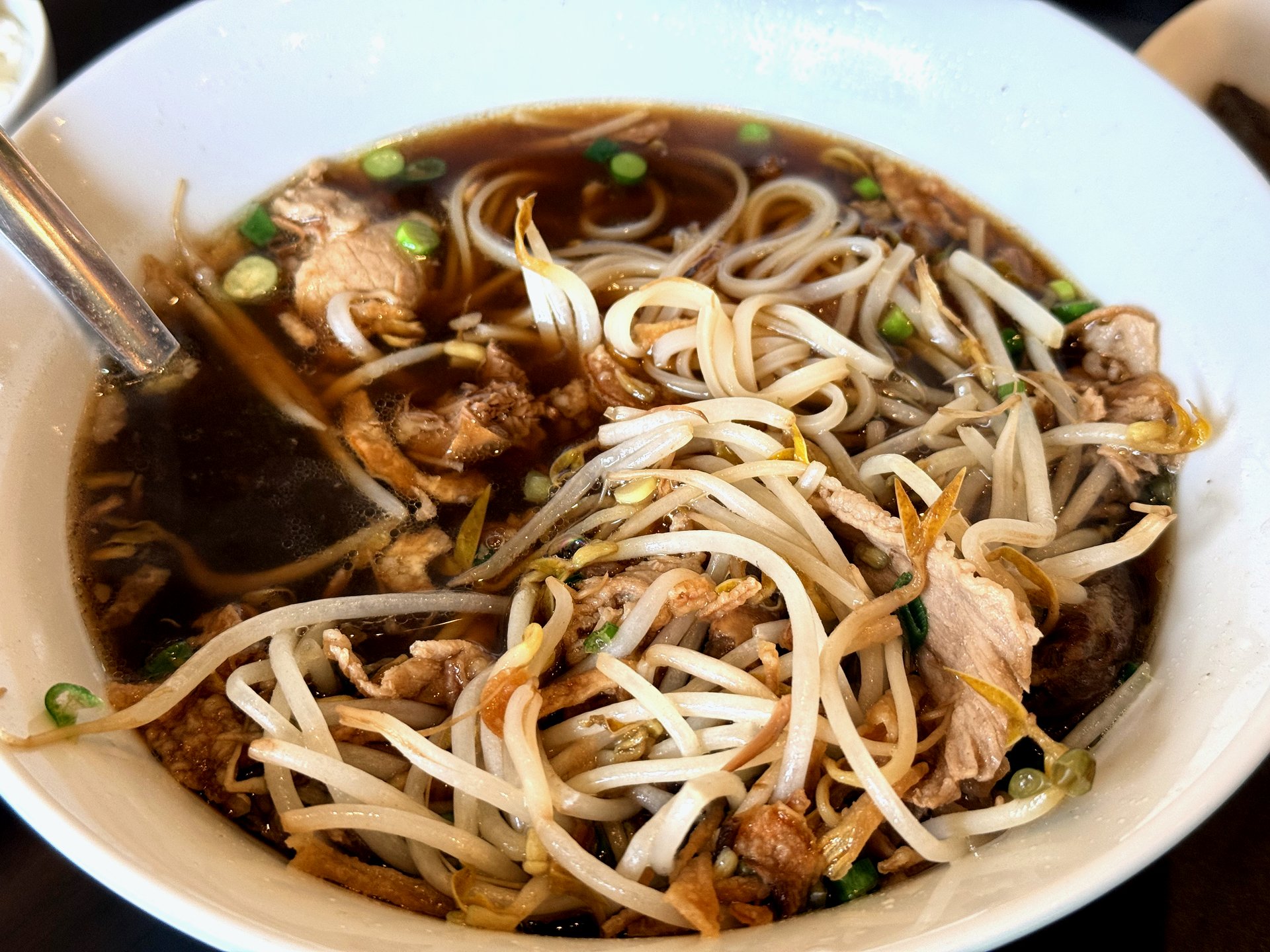 Meat-lover Thai rice noodle soup at Pai Deng Yeongjongdo - a white bowl of dark, clear brown broth with rice noodles, bean sprouts, thin slices of boiled beef and galbi, and chopped scallions, a spoon resting in the bowl