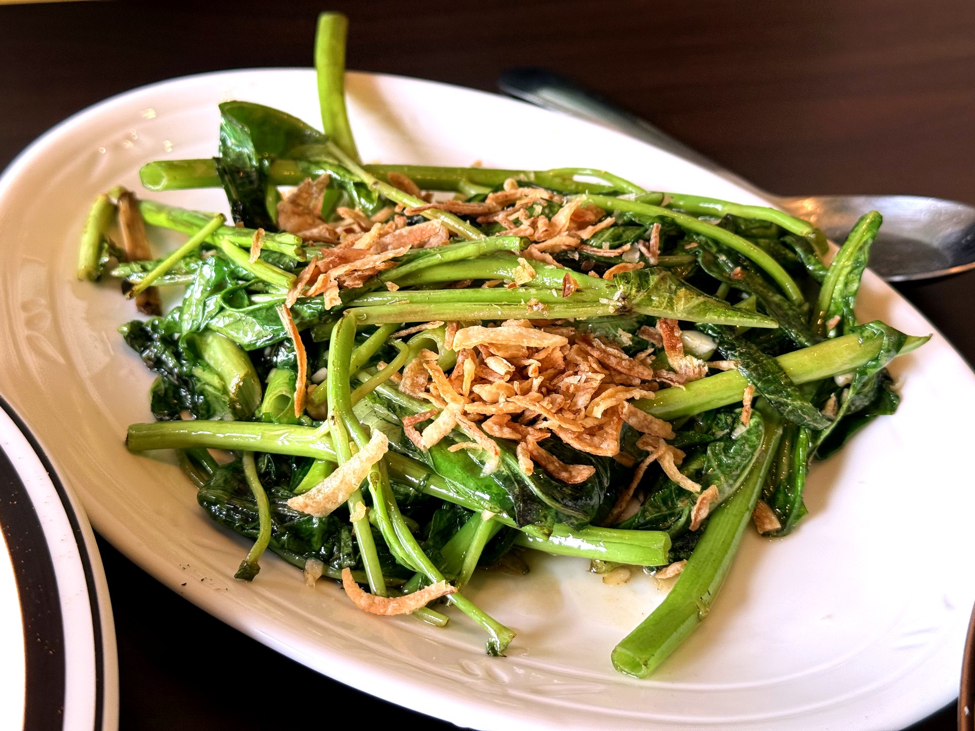 Morning glory stir-fry at Pai Deng Yeongjongdo - a white oval plate of bright green stir-fried water spinach with long snappy stems, glossy with oil and garlic and topped with golden crispy fried shallots
