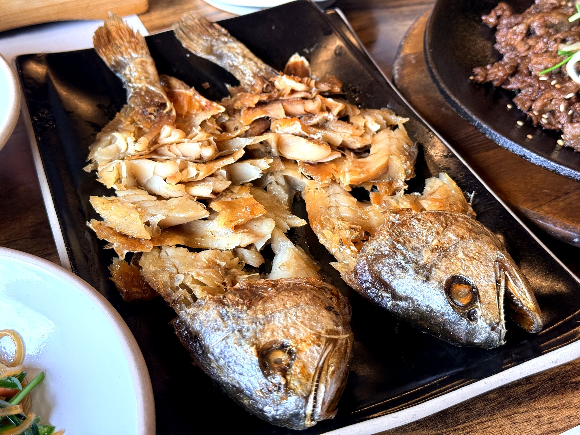 Haenuri Dongsan Deulbap 보리굴비 (dried salted croaker) - two whole grilled golden-brown croaker fish on a black rectangular plate, the flesh expertly flaked off the bone into savory pieces, a Korean delicacy