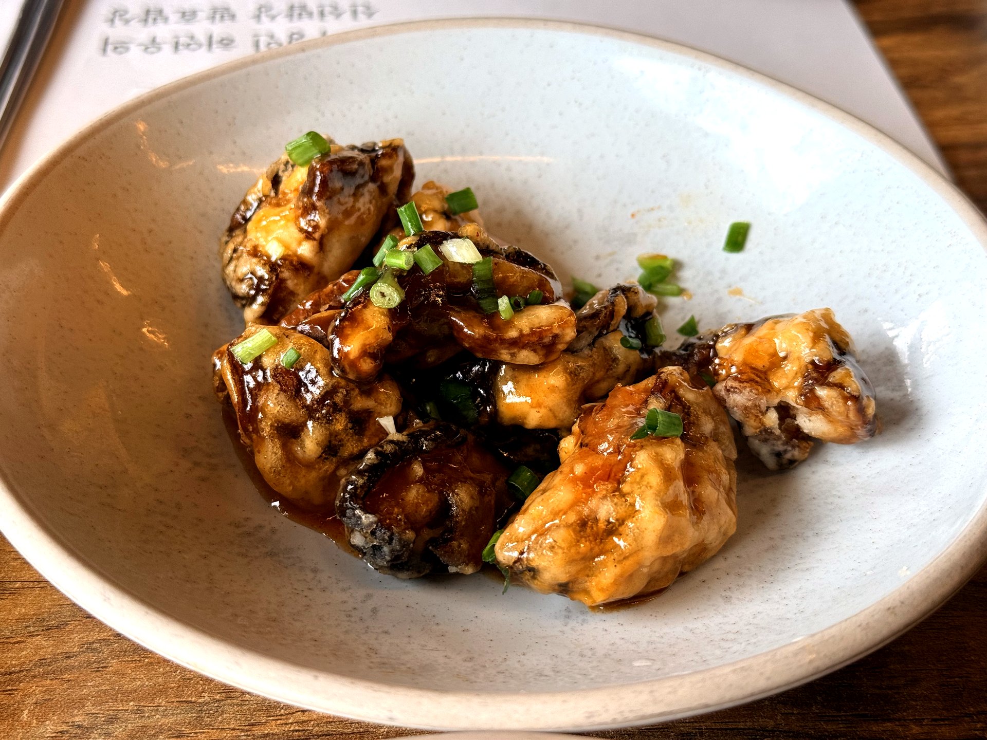 Haenuri Dongsan Deulbap battered fried oysters in a light savory glaze, garnished with chopped green onion, in a cream ceramic bowl - a side dish in the Korean set meal