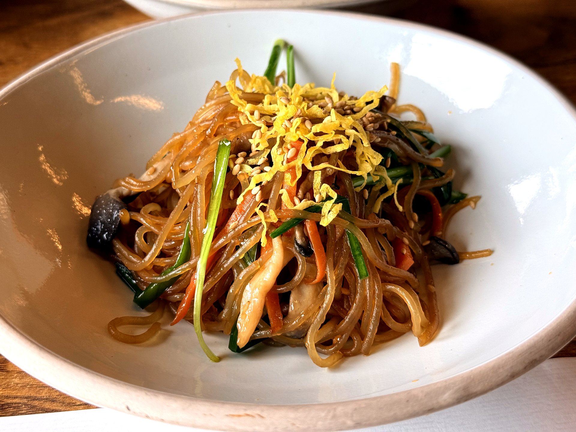 Haenuri Dongsan Deulbap 잡채 (japchae) - glossy stir-fried sweet-potato glass noodles tossed with mushrooms, carrot, spinach and chive, topped with fried egg strips and sesame, in a white oval bowl