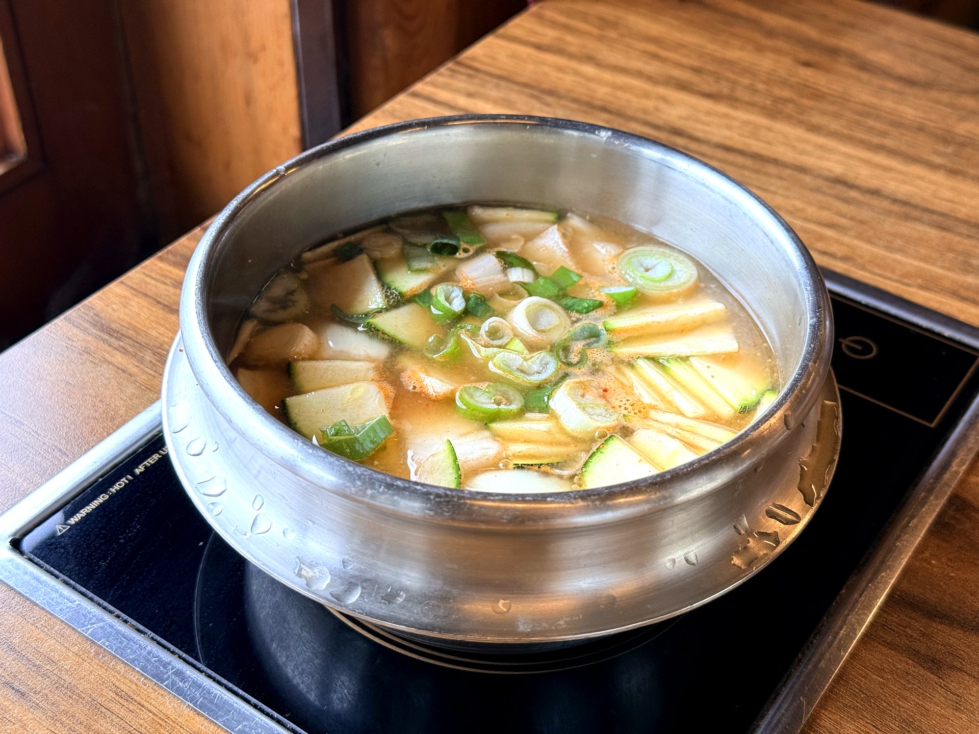 Haenuri Dongsan Deulbap 된장찌개 (soybean-paste stew) - a bubbling metal pot of Korean doenjang stew with zucchini, radish and green onion, simmering on a tabletop induction burner