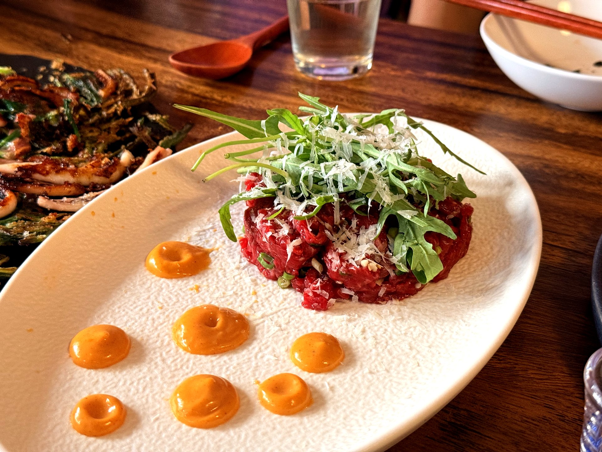 Fusion yukhoe at Jurak in Mullae, Seoul - Korean seasoned raw beef tartare piled on a white plate, topped with a tangle of fresh arugula and finely grated parmesan cheese, with dots of an orange savory sauce around the plate
