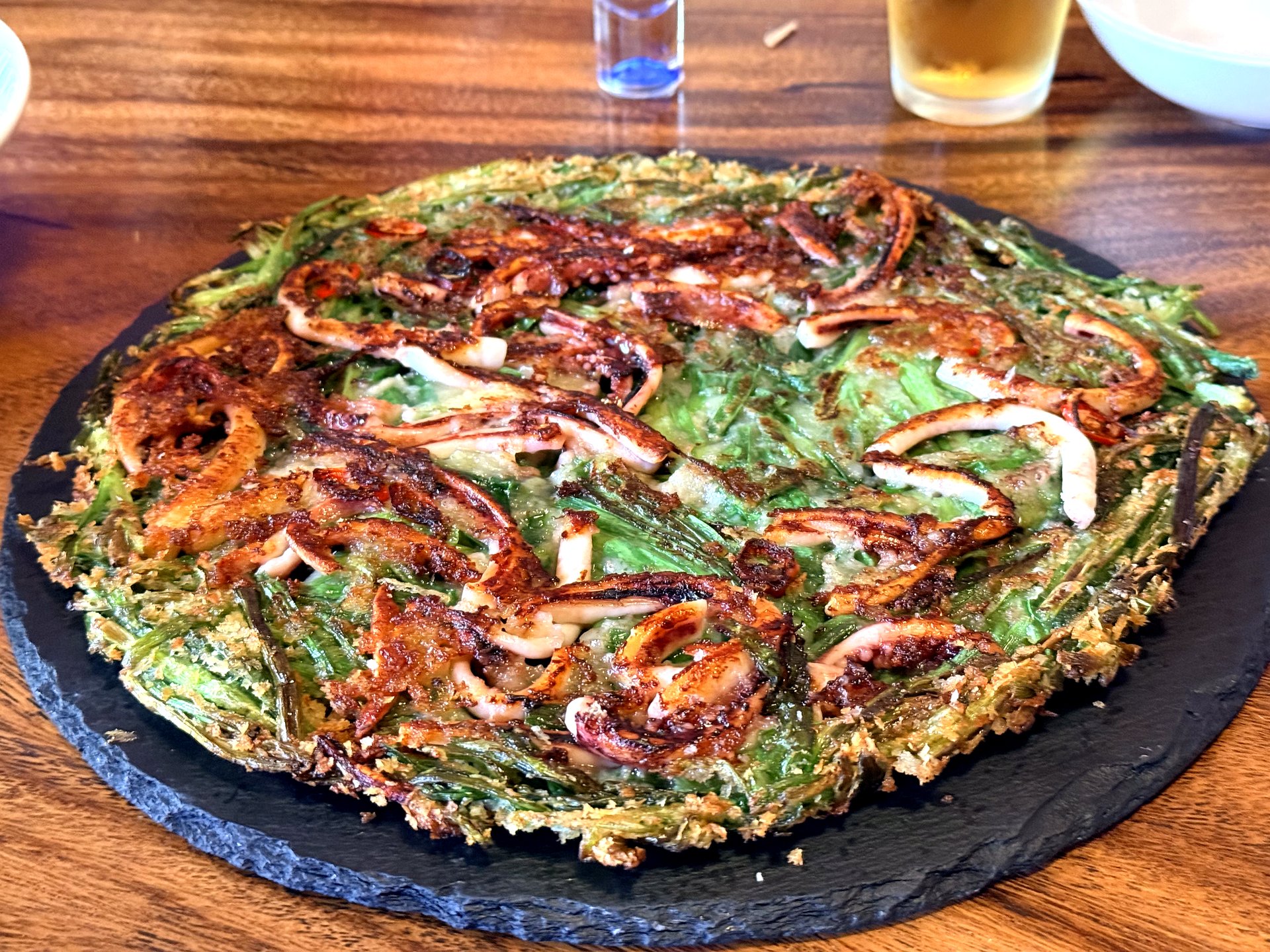 Squid and minari jeon at Jurak in Mullae, Seoul - a large round crispy Korean pancake made with water-dropwort greens and topped with charred sliced squid, served on a black slate plate with a shot glass of clear liquor beside it