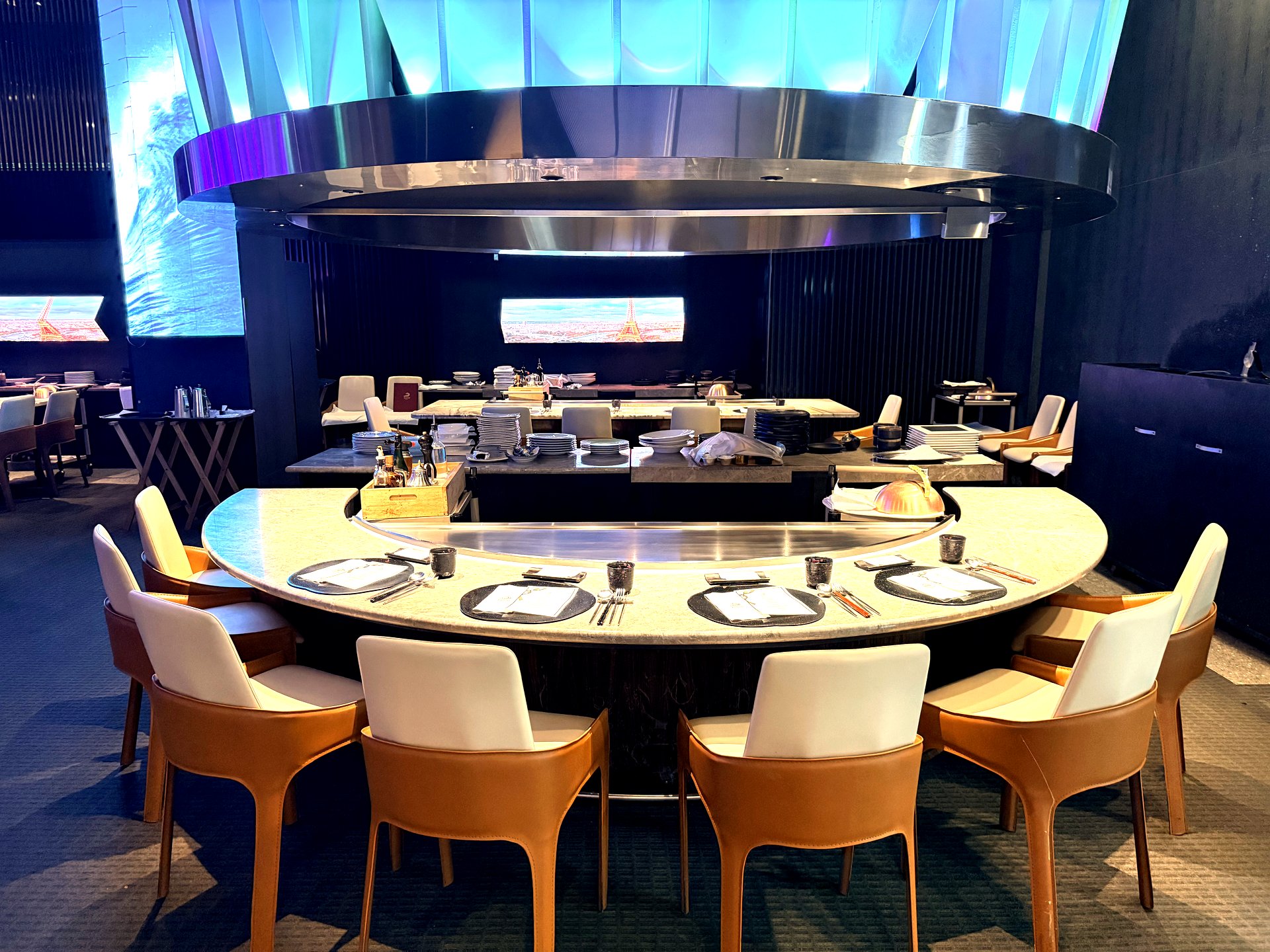 Interior of Kawabe Teppanyaki at The Riverside Hotel Seoul - a dramatic circular teppanyaki counter with a large polished steel hood, blue and purple mood lighting, tan leather chairs and marble counters set with black plates