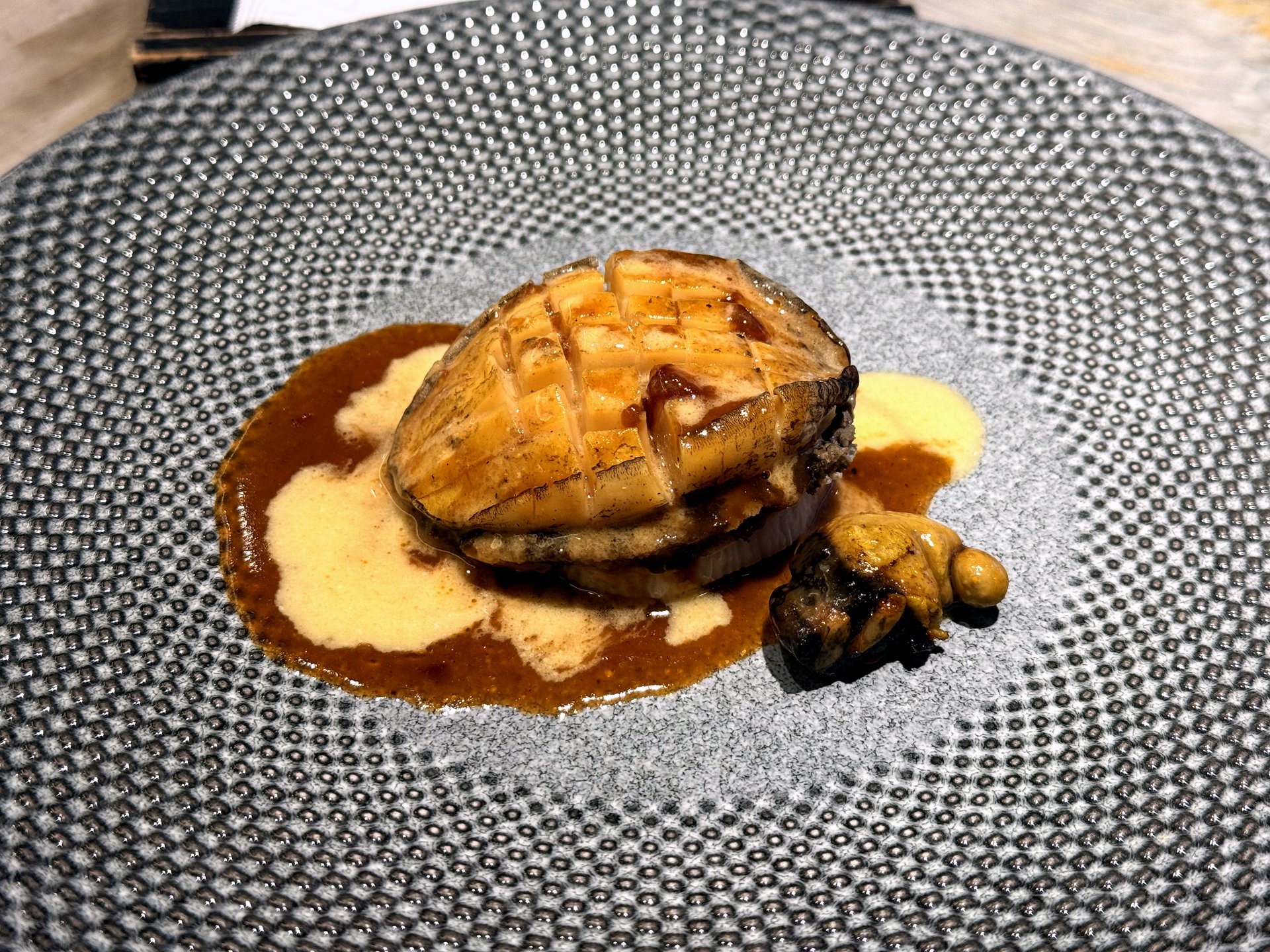 Course five at Kawabe Teppanyaki Seoul - a large scored rock-salt abalone glazed and plated on a speckled grey plate over a pool of sauce, with the abalone liver alongside