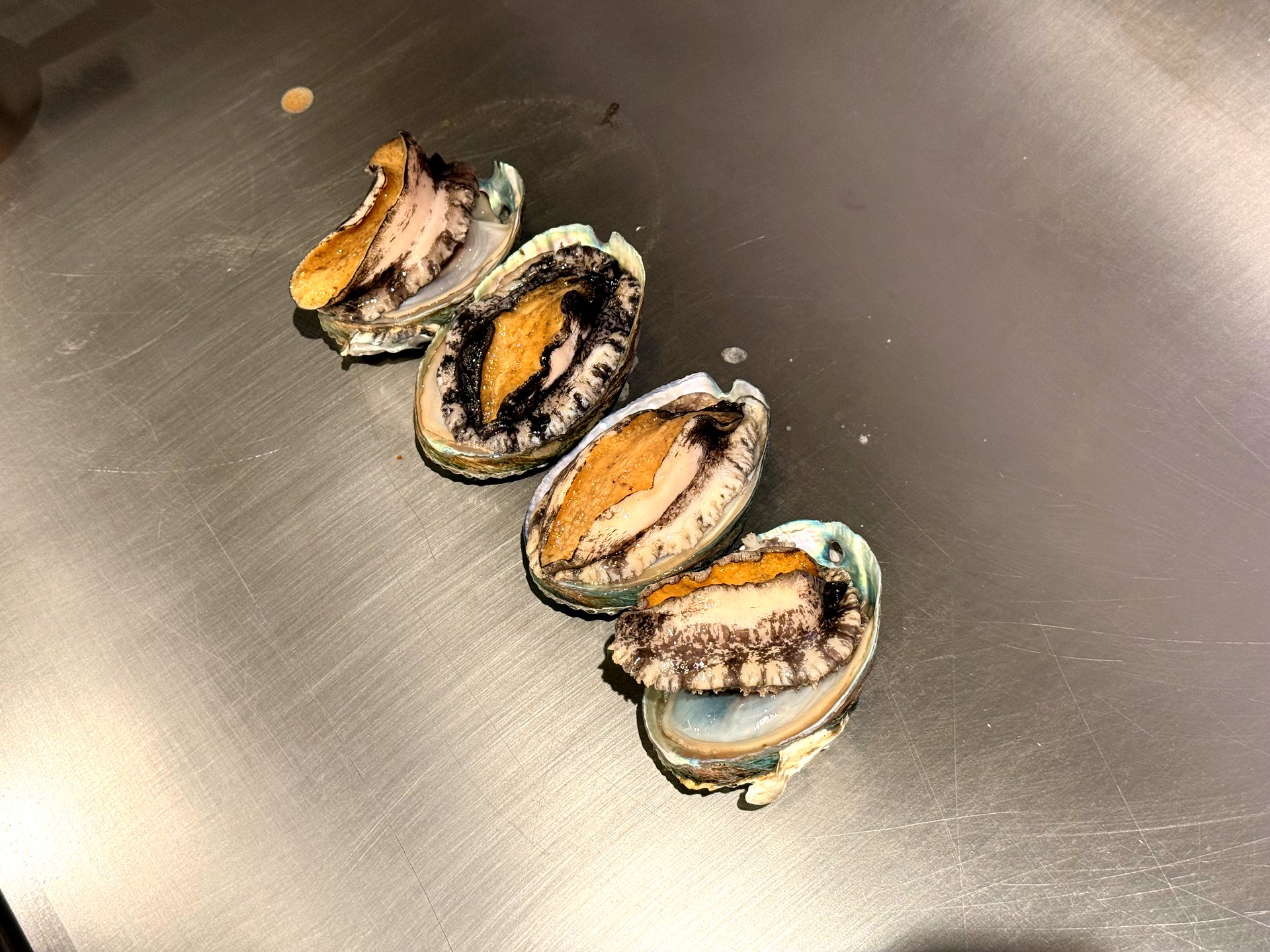 Four live abalones in their shells laid out on the teppanyaki griddle at Kawabe Teppanyaki Seoul before cooking, showing the orange-brown abalone flesh and iridescent shells
