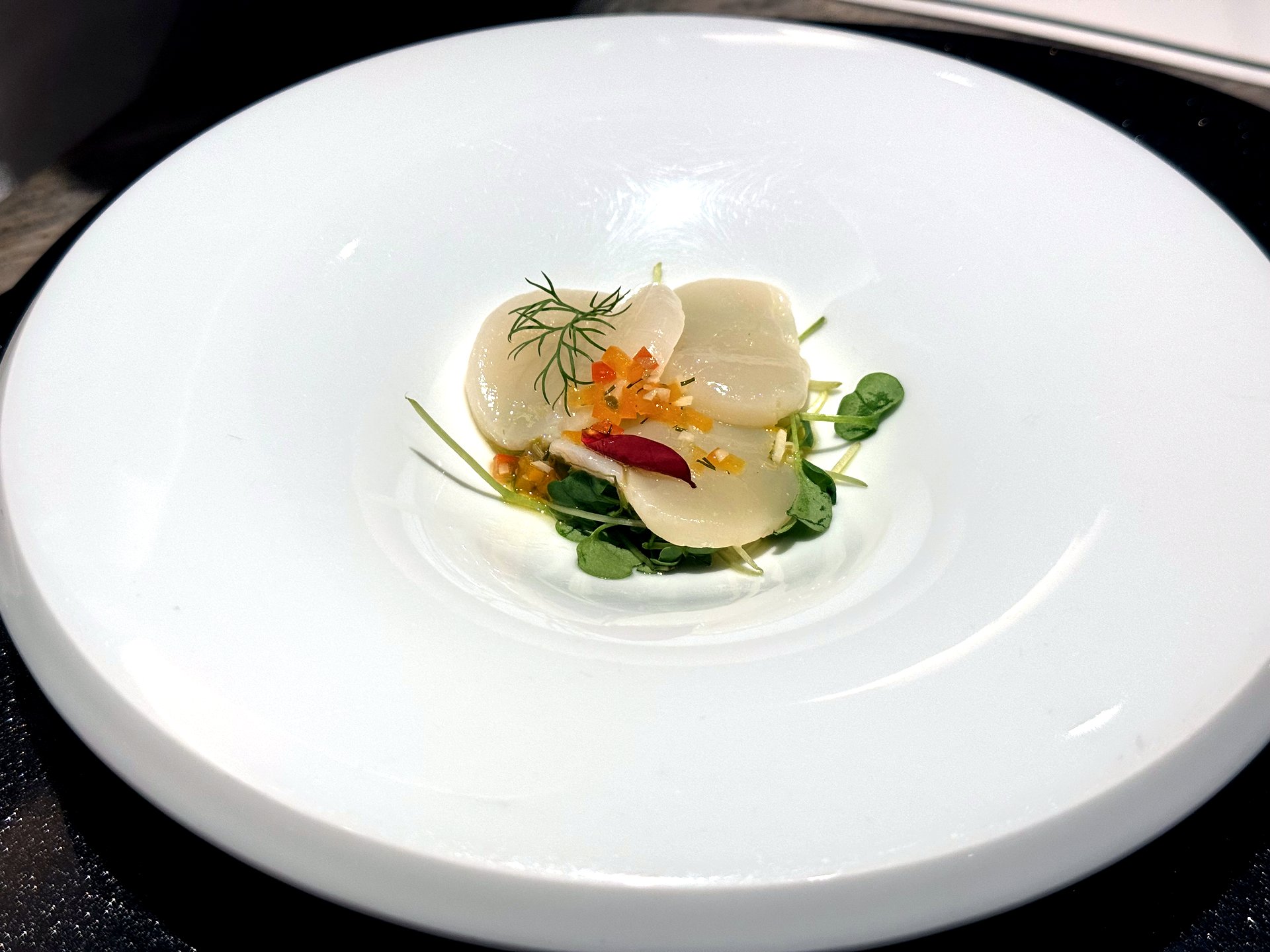 Course one at Kawabe Teppanyaki Seoul - an elegant chilled scallop appetizer, slices of raw scallop fanned over micro-greens with a citrus-chili dressing and a shiso leaf, on a white bowl