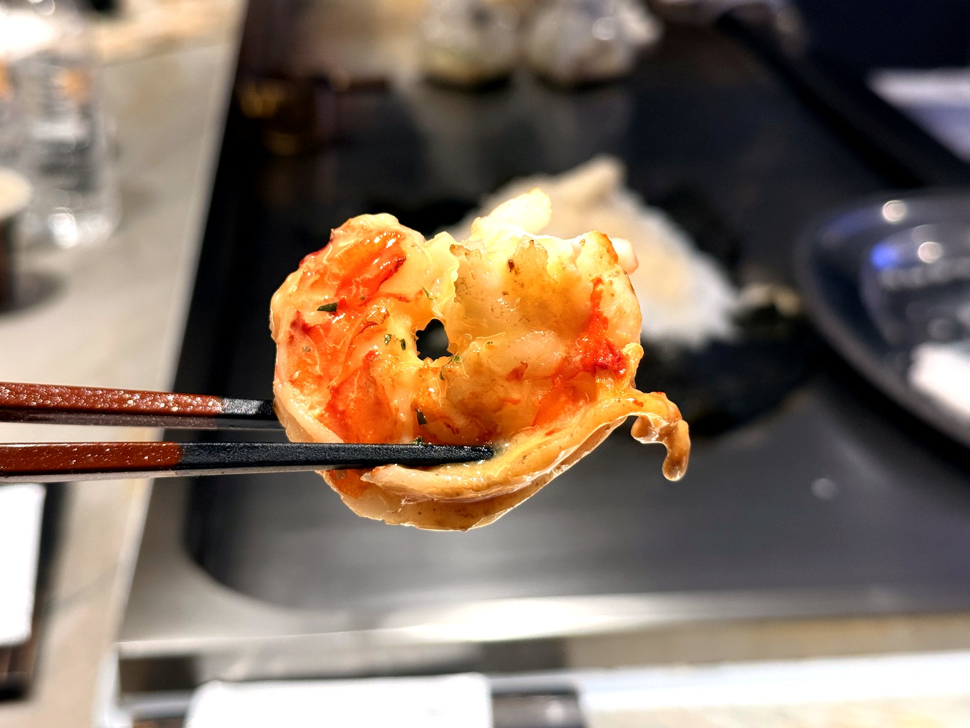 Course three at Kawabe Teppanyaki Seoul - a close-up of a glossy grilled black tiger prawn held in chopsticks, its shell lightly charred and flesh juicy