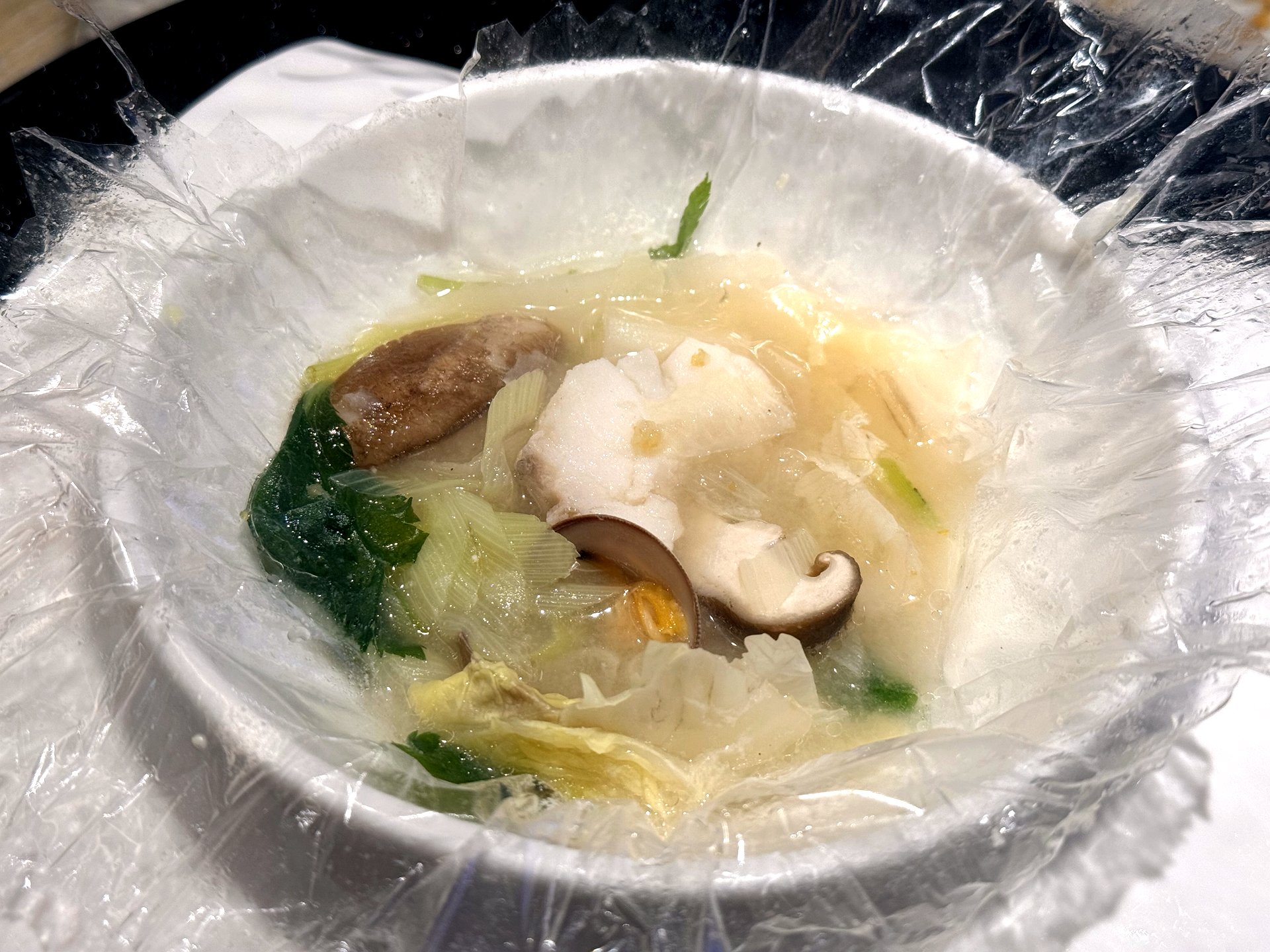 The seafood papillote opened at Kawabe Teppanyaki Seoul - a peeled-back edible-film pouch holding a clear milky broth with scallop, shiitake mushroom, leek and green vegetables