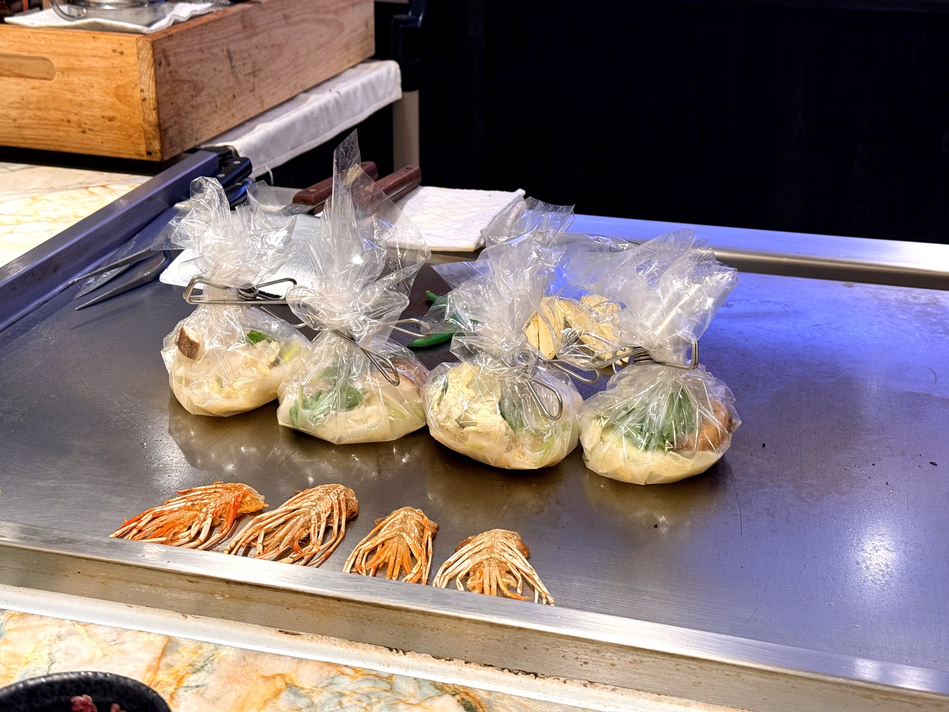 Seafood papillote cooking at Kawabe Teppanyaki Seoul - four clear edible-film pouches twisted and clipped shut like candy wrappers, filled with seafood and vegetables, steaming on the steel griddle beside prawns