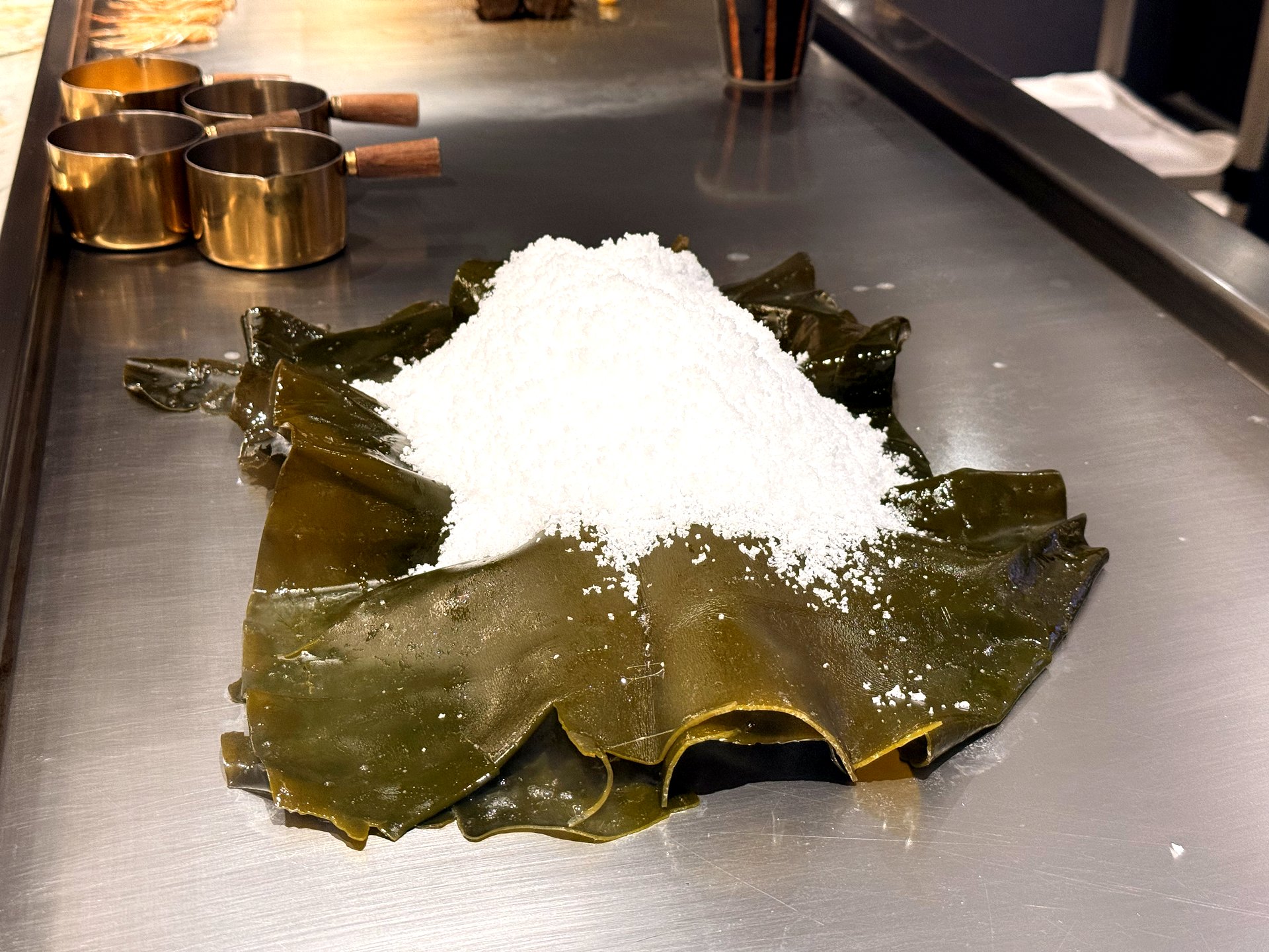Rock-salt abalone being slow-cooked at Kawabe Teppanyaki Seoul - a large mound of coarse white salt piled over sheets of dark green kelp on the steel griddle, with brass sauce pots nearby