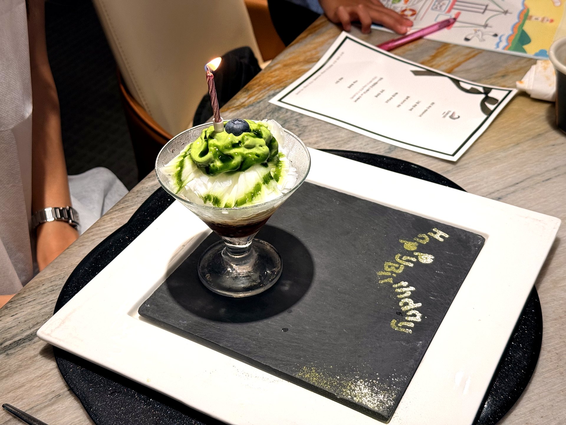 Course eight at Kawabe Teppanyaki Seoul - a matcha-cream shaved-ice bingsu in a coupe glass topped with a blueberry and a lit candle, on a white plate over a black slate hand-lettered with a gold 'Happy Birthday'