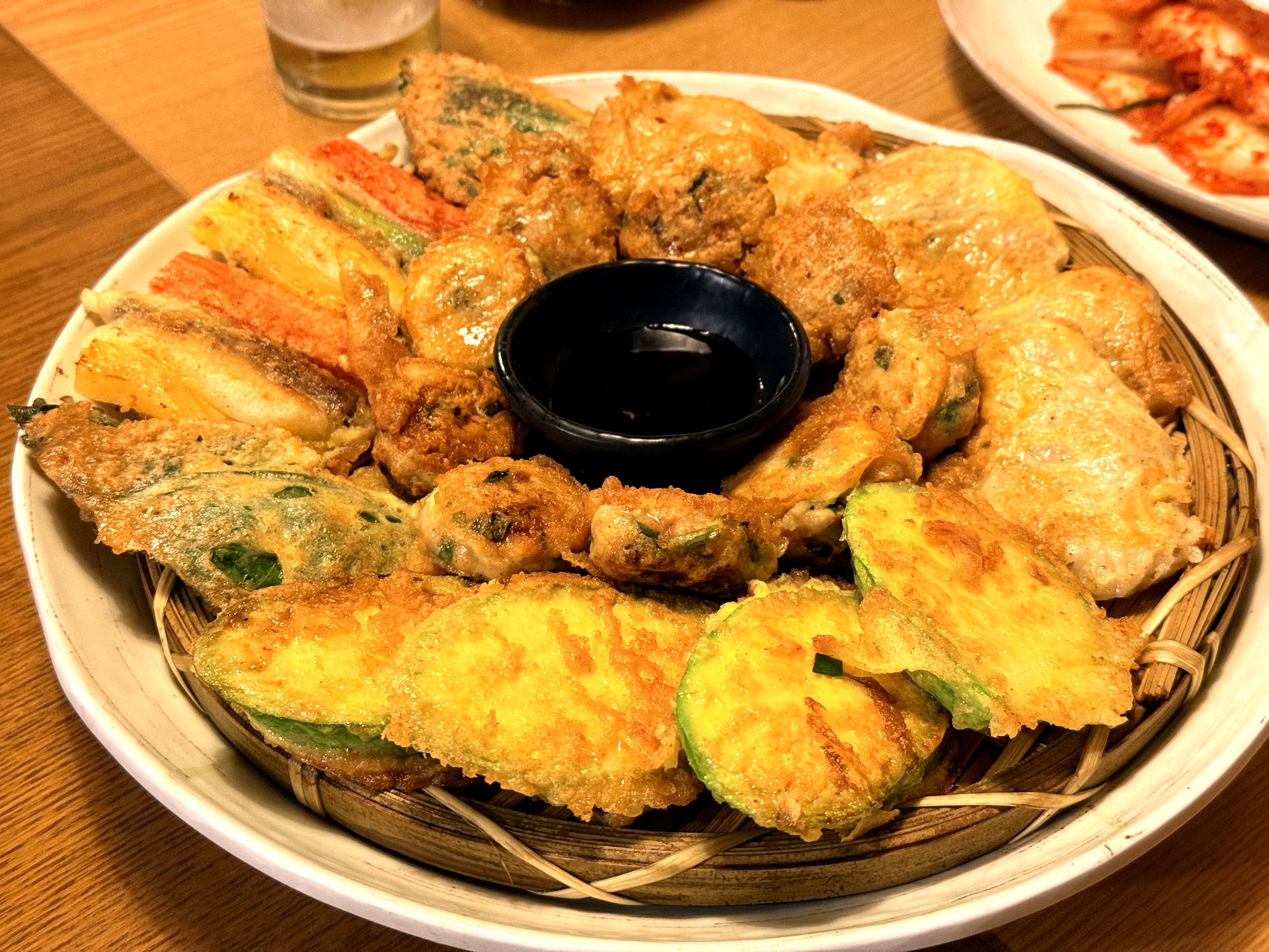 Mapo Naru 모듬전 (assorted jeon platter) - a bamboo basket piled with golden pan-fried Korean savory pancakes including zucchini jeon, fish jeon, meat patties and crab stick, arranged in a fan around a small dish of dark soy dipping sauce, with kimchi on a white plate in the background