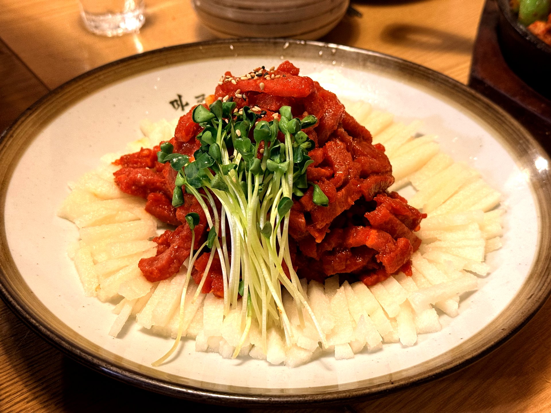 Mapo Naru 육회 (Korean beef tartare) - a mound of glossy red hand-cut seasoned raw beef on a bed of julienned white Korean pear and radish, topped with fresh green microgreens and sesame seeds, on a cream ceramic plate branded with the restaurant name