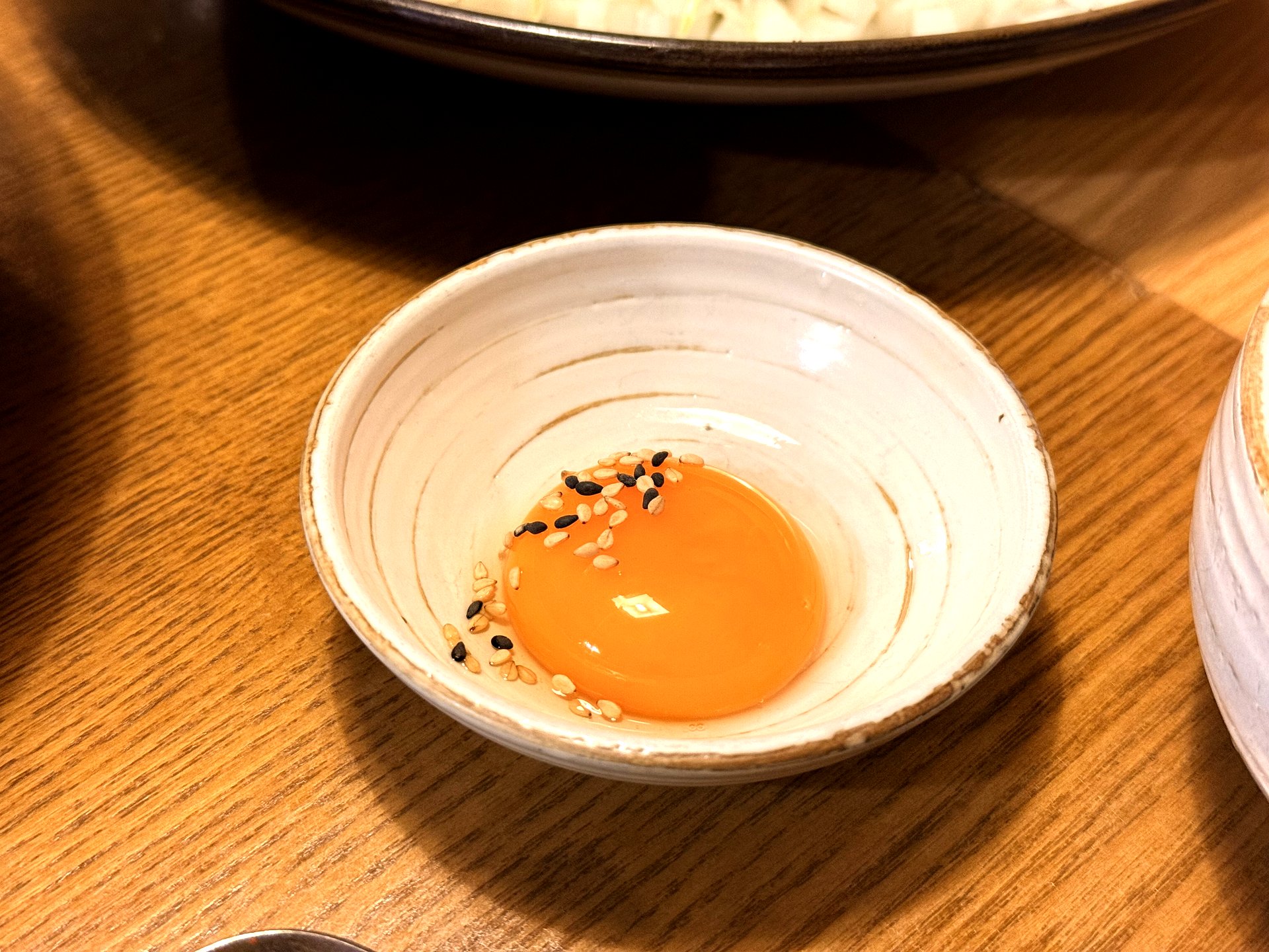 A single glossy orange raw egg yolk in a small white ceramic dish sprinkled with black and white sesame seeds, the traditional accompaniment to Korean yukhoe beef tartare, on a wooden table at Mapo Naru