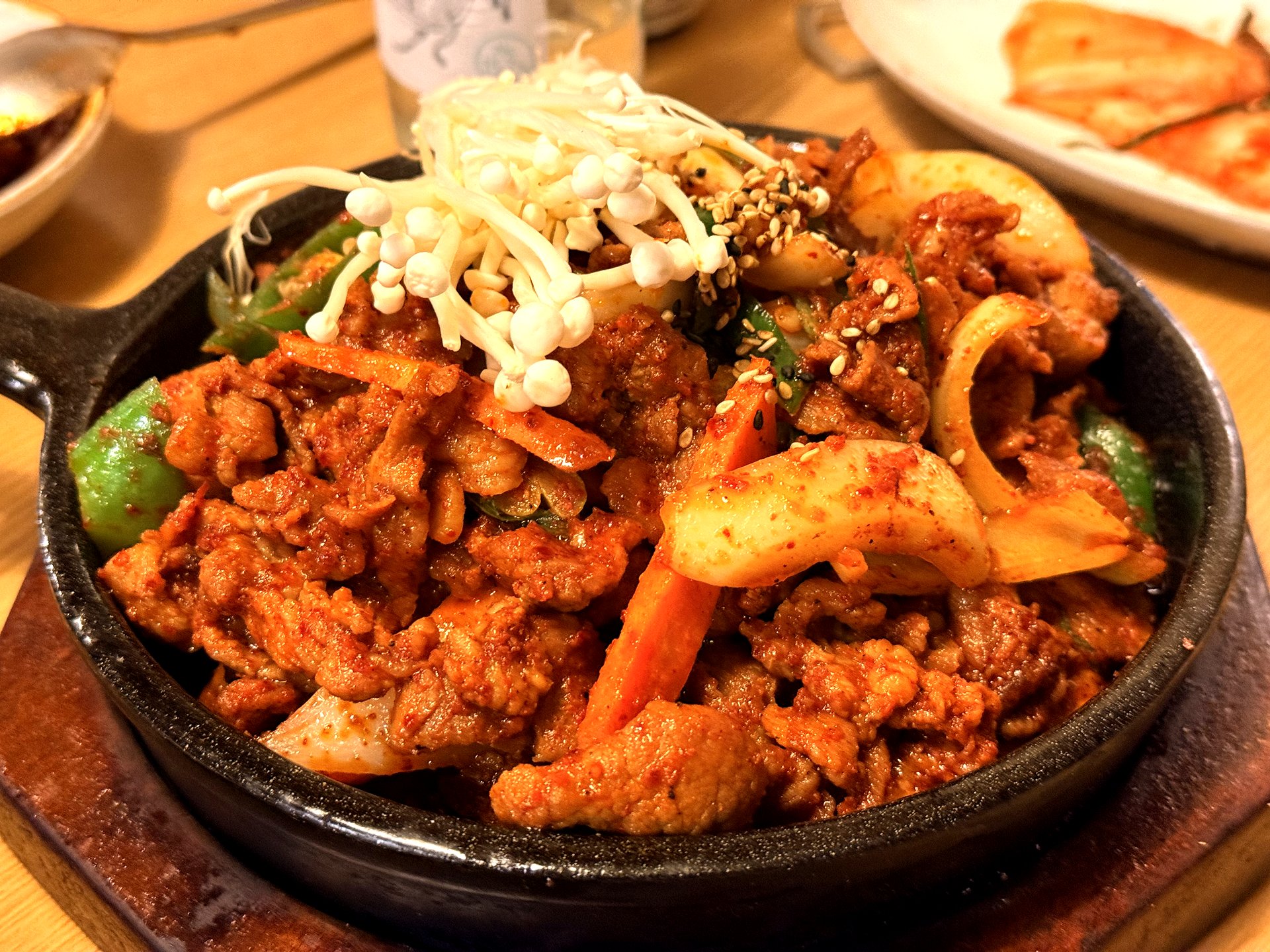 Mapo Naru 제육볶음 (spicy pork stir-fry) - thin slices of pork glazed in red gochujang sauce with onion, green pepper and carrot, sizzling on a black cast-iron pan and topped with a nest of fresh white enoki mushrooms and sesame seeds