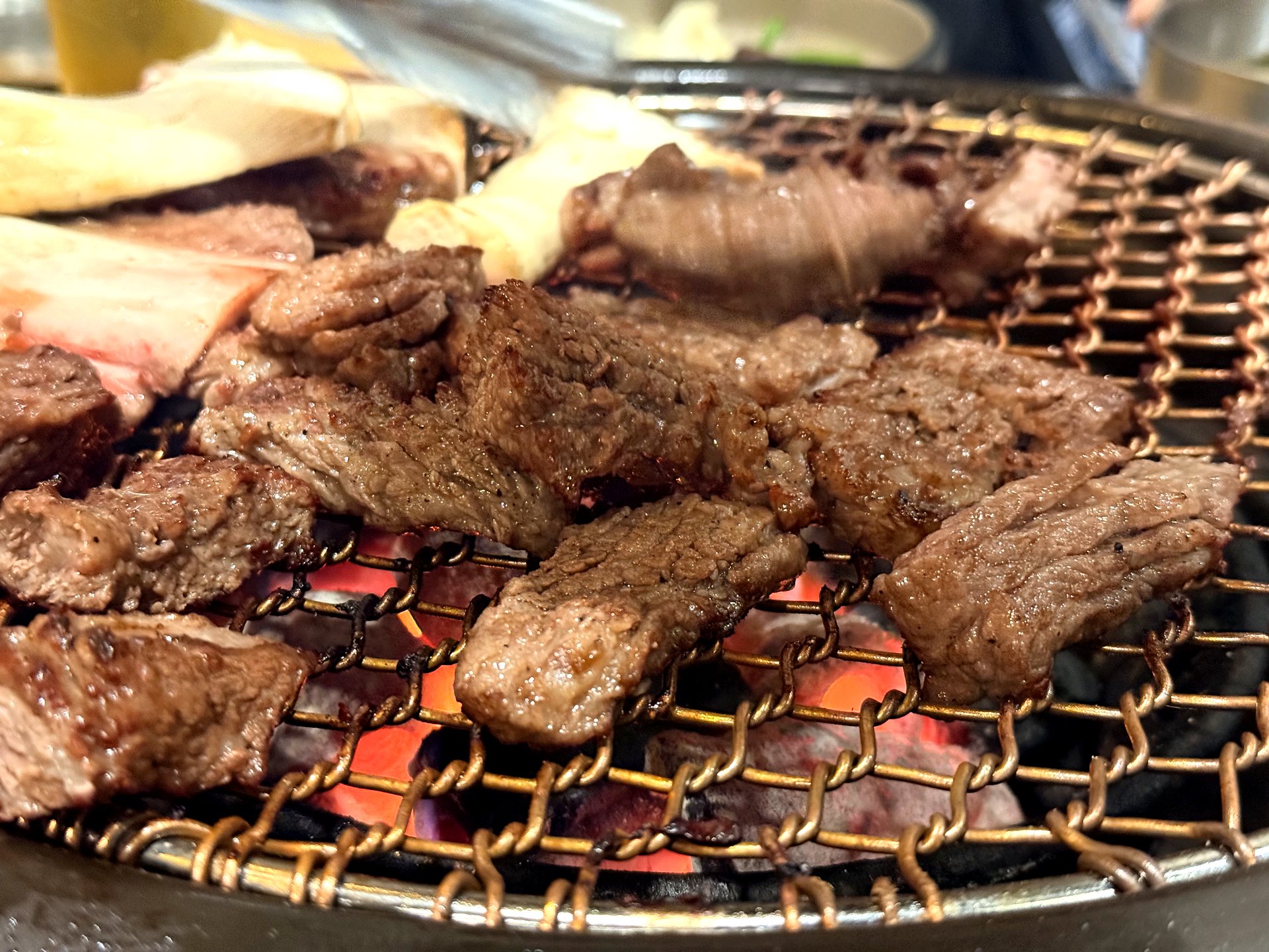 Beef galbi grilling over glowing red charcoal on a copper-mesh grill at Oso Galbi - pieces of marinated and marbled beef short rib browning and caramelizing, with the orange glow of charcoal visible through the mesh