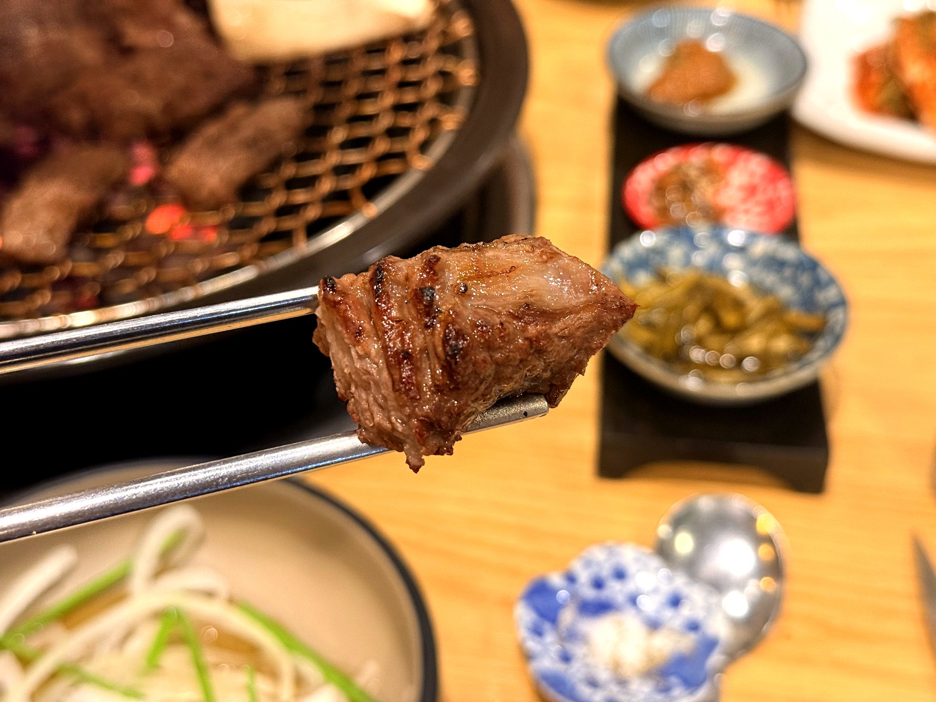 A perfectly grilled bite-sized piece of beef galbi held up in metal tongs at Oso Galbi, charred and glistening on the outside and juicy inside, with the charcoal grill and banchan dishes blurred in the background