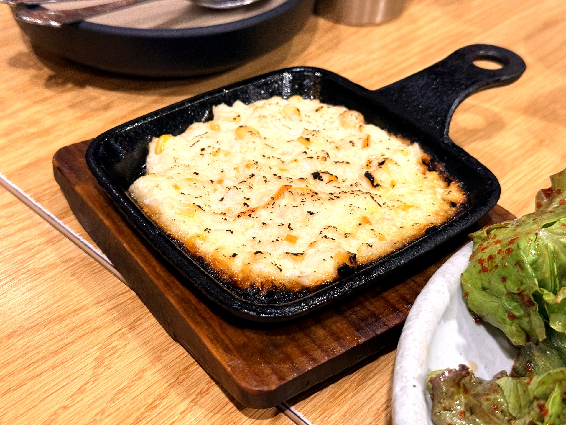 Oso Galbi 콘치즈 (corn cheese) - sweet corn kernels baked in bubbling golden melted cheese in a small black cast-iron skillet on a wooden board, a popular Korean BBQ side dish