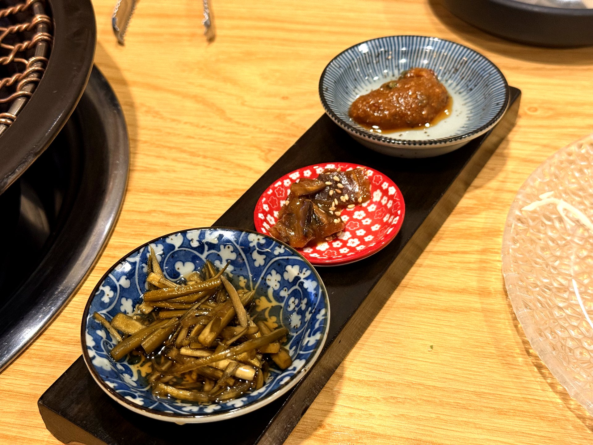 Oso Galbi banchan trio on a black tray - three small patterned ceramic dishes holding soy-braised burdock strips, marinated vegetables, and a savory seasoned paste, served beside the charcoal grill at a Korean BBQ restaurant