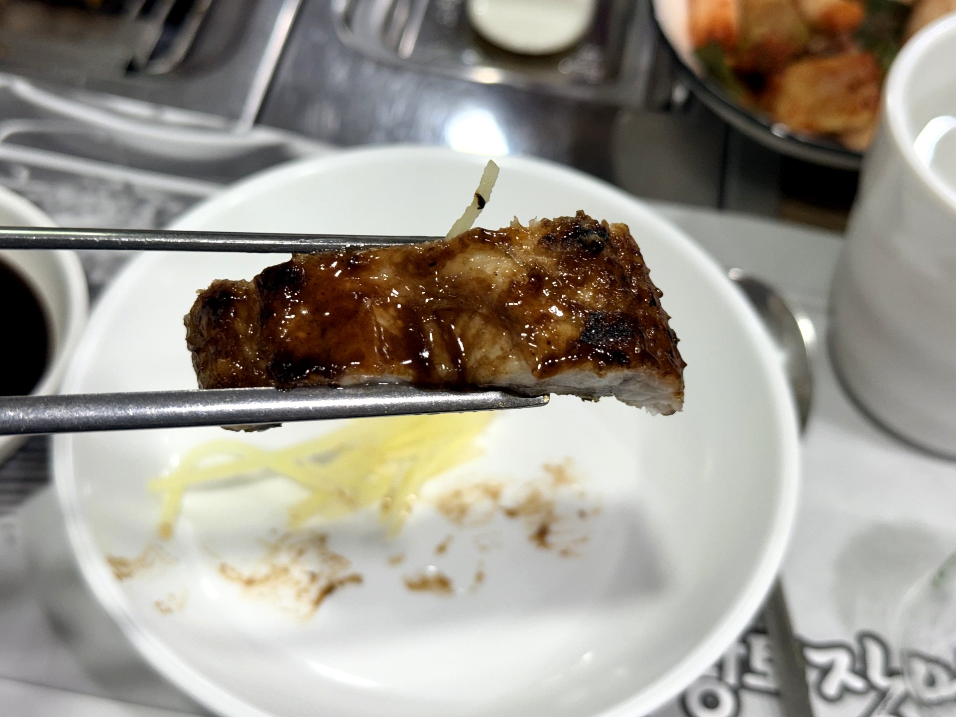 A close-up of a single glazed piece of grilled freshwater eel held in metal chopsticks at Hwangto Jangeo Maeul, glossy and caramelized on the outside with tender white flesh, above a plate with slivers of pickled ginger