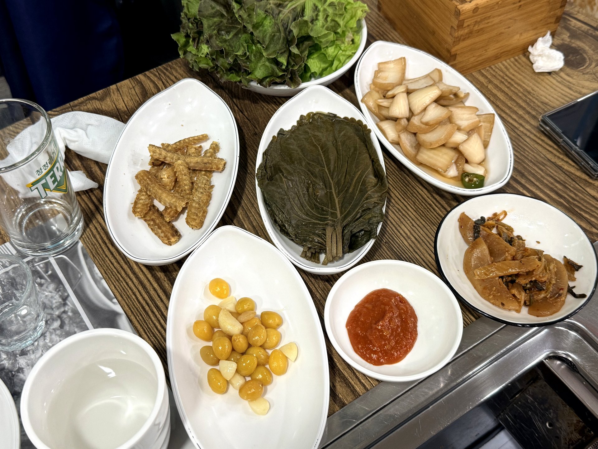Hwangto Jangeo Maeul ssam and banchan spread - a bowl of fresh lettuce leaves, a plate of pickled perilla leaves, pickled radish and onion, crispy fried burdock sticks, ginkgo nuts with garlic, and a small dish of red chili-vinegar sauce, arranged on a wooden table