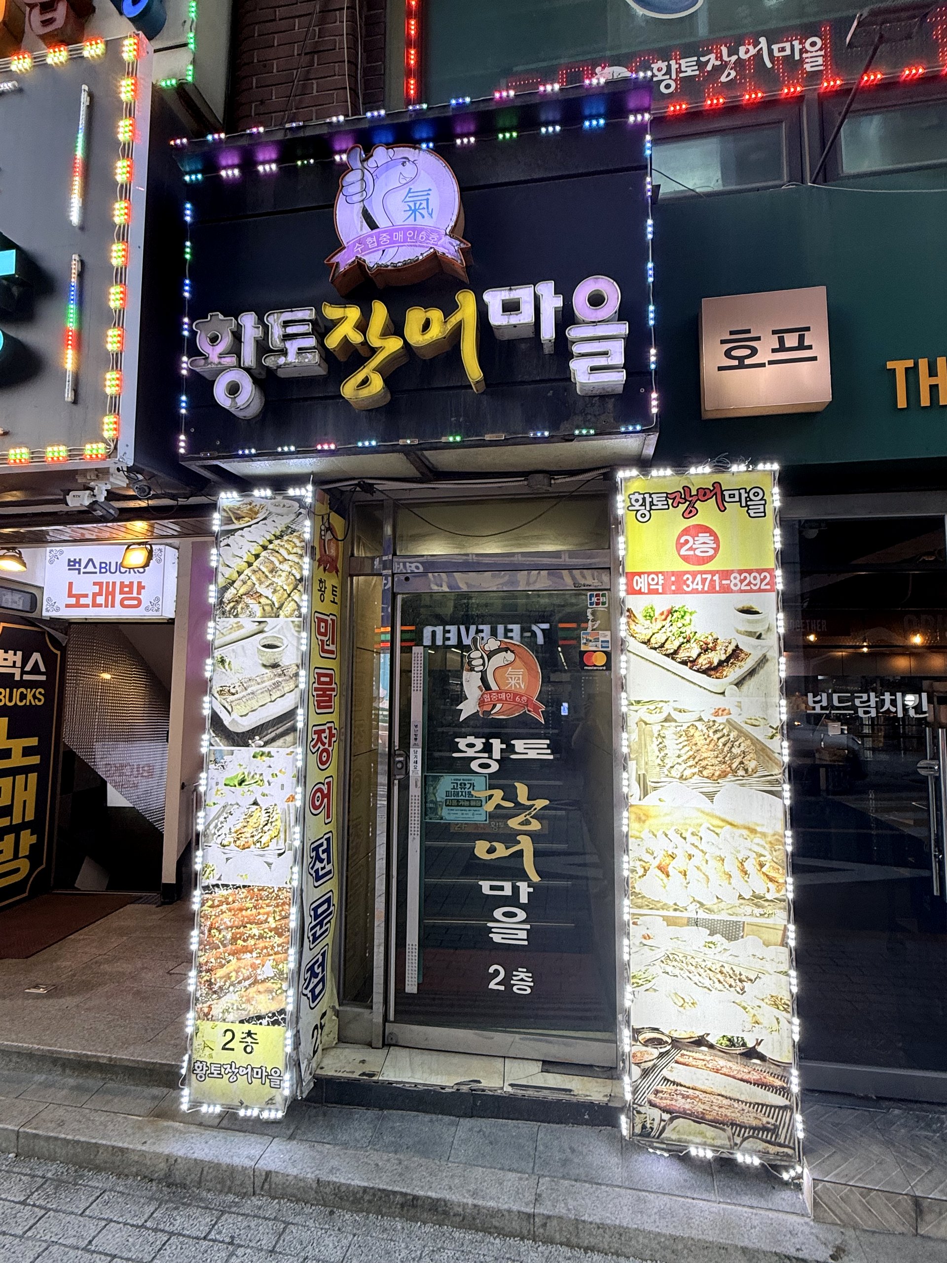 Hwangto Jangeo Maeul storefront at night - a brightly lit second-floor Korean eel restaurant sign reading 황토장어마을 (with a cartoon eel mascot giving a thumbs-up) and 2층, surrounded by LED lights and photo panels of grilled eel dishes, near Sadang Station Seoul
