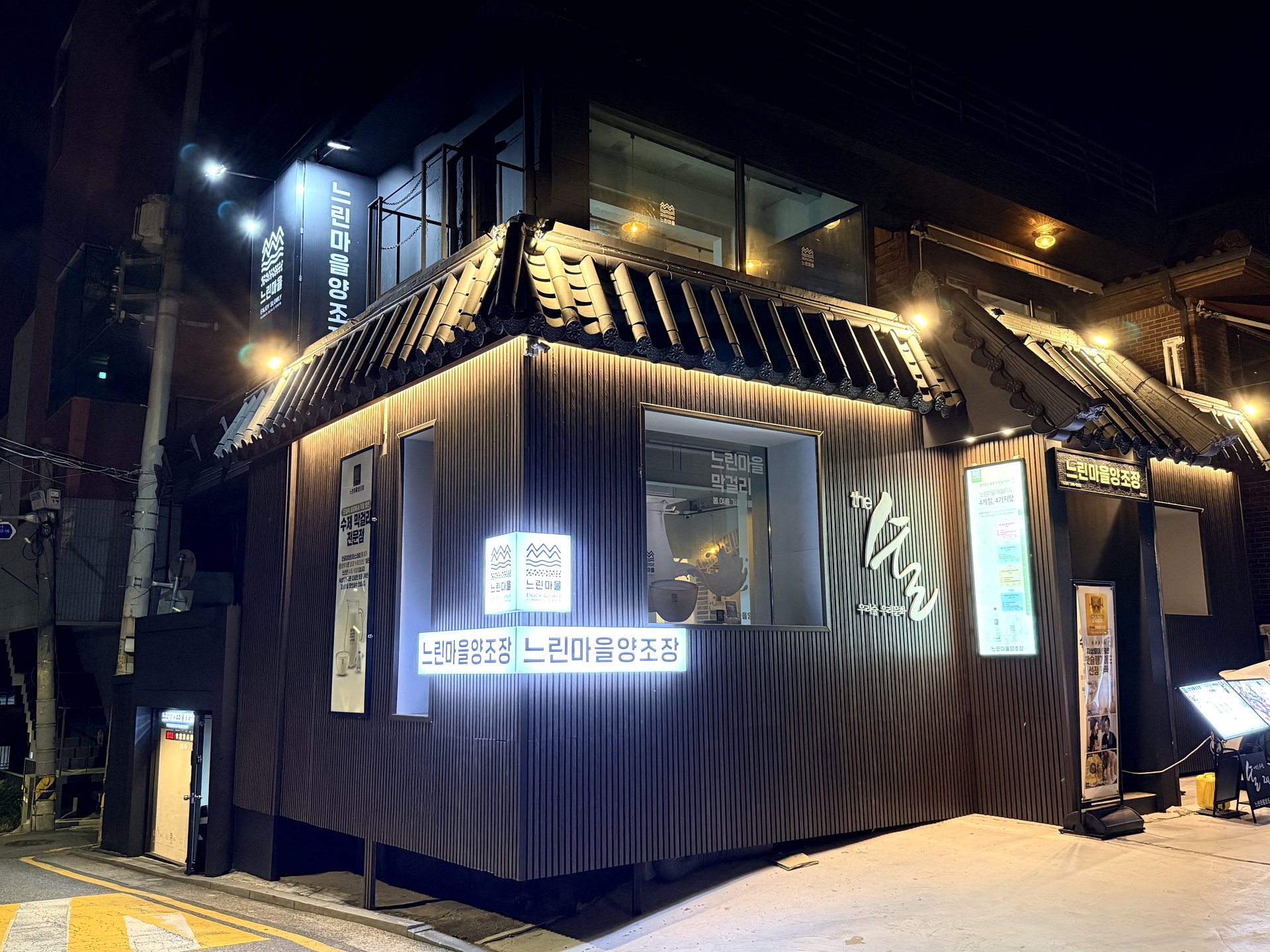 Neurin Maeul Brewery Hakdong exterior at night - a modern two-story hanok-style building with a traditional grey tiled roof and dark vertical timber cladding, warmly lit, with illuminated 느린마을양조장 (Neurin Maeul Brewery) signs on the corner in Gangnam, Seoul