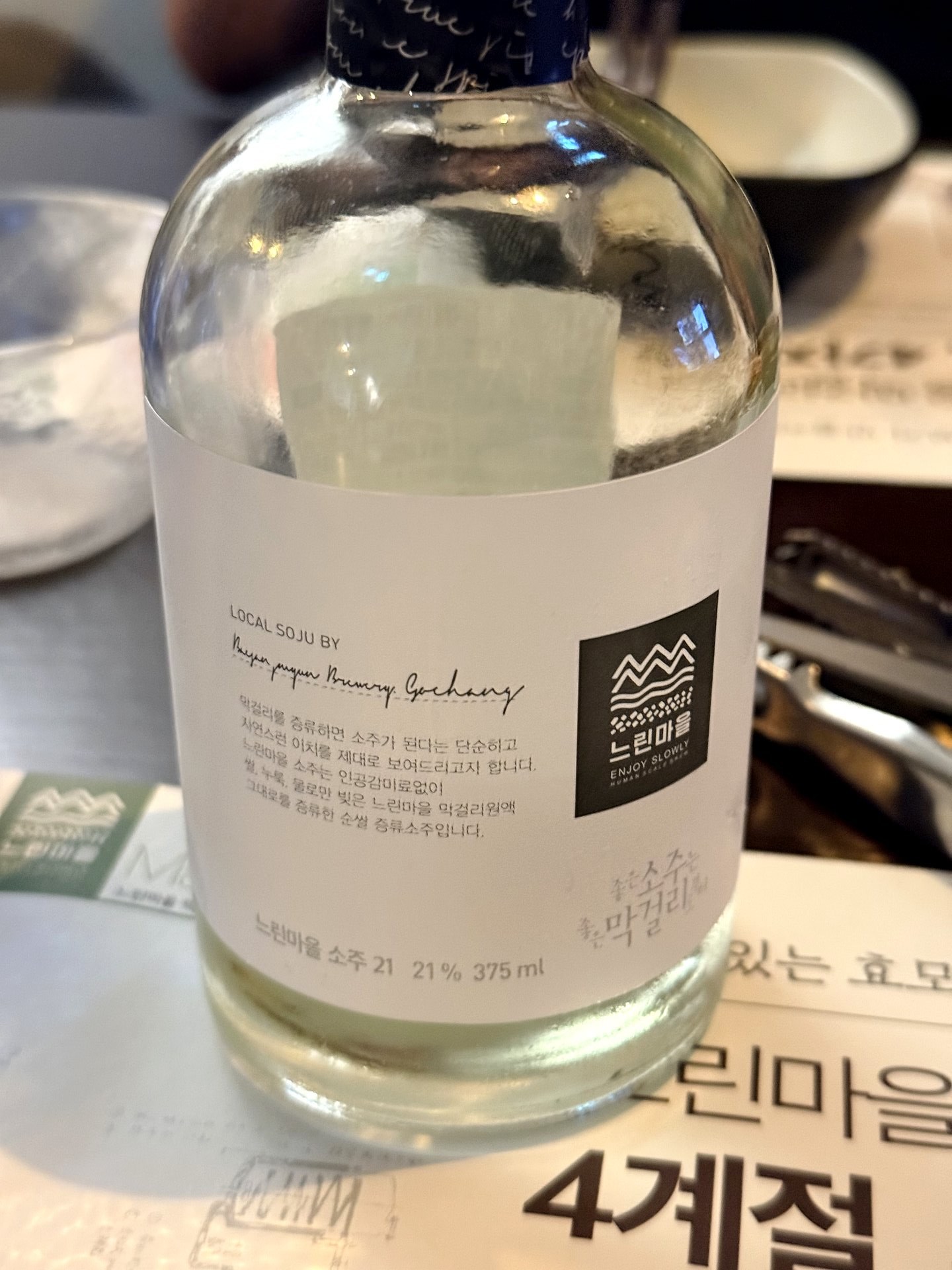 A 375ml bottle of 느린마을 소주 21 (Neurin Maeul distilled soju, 21% ABV) lying on its side, a clear premium Korean craft soju with a minimalist white label reading LOCAL SOJU, on a table at the brewery-pub