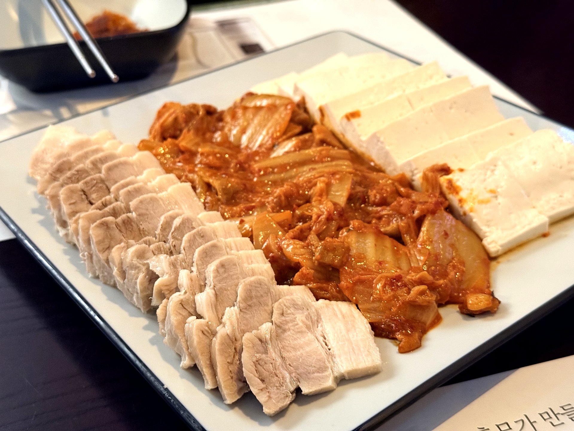 Neurin Maeul Brewery 수육 두부김치 - a long white plate with rows of thinly sliced boiled pork belly (suyuk) on the left, a mound of stir-fried red kimchi in the middle, and slices of white tofu on the right