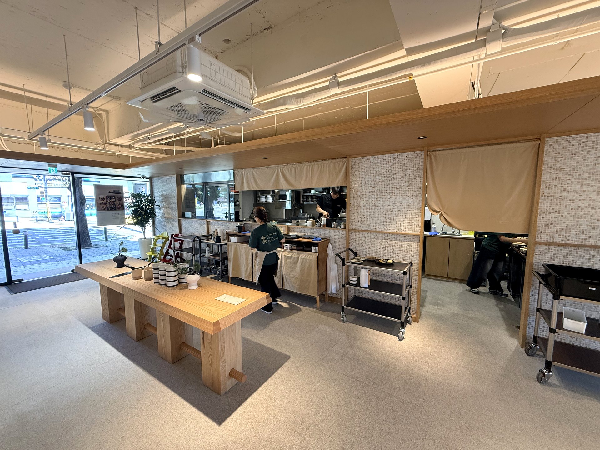 The open kitchen at Ondang - staff in dark green KOREAN TOFU CULTURE aprons working behind a counter with linen curtains, preparing fresh tofu, in a bright modern Korean restaurant