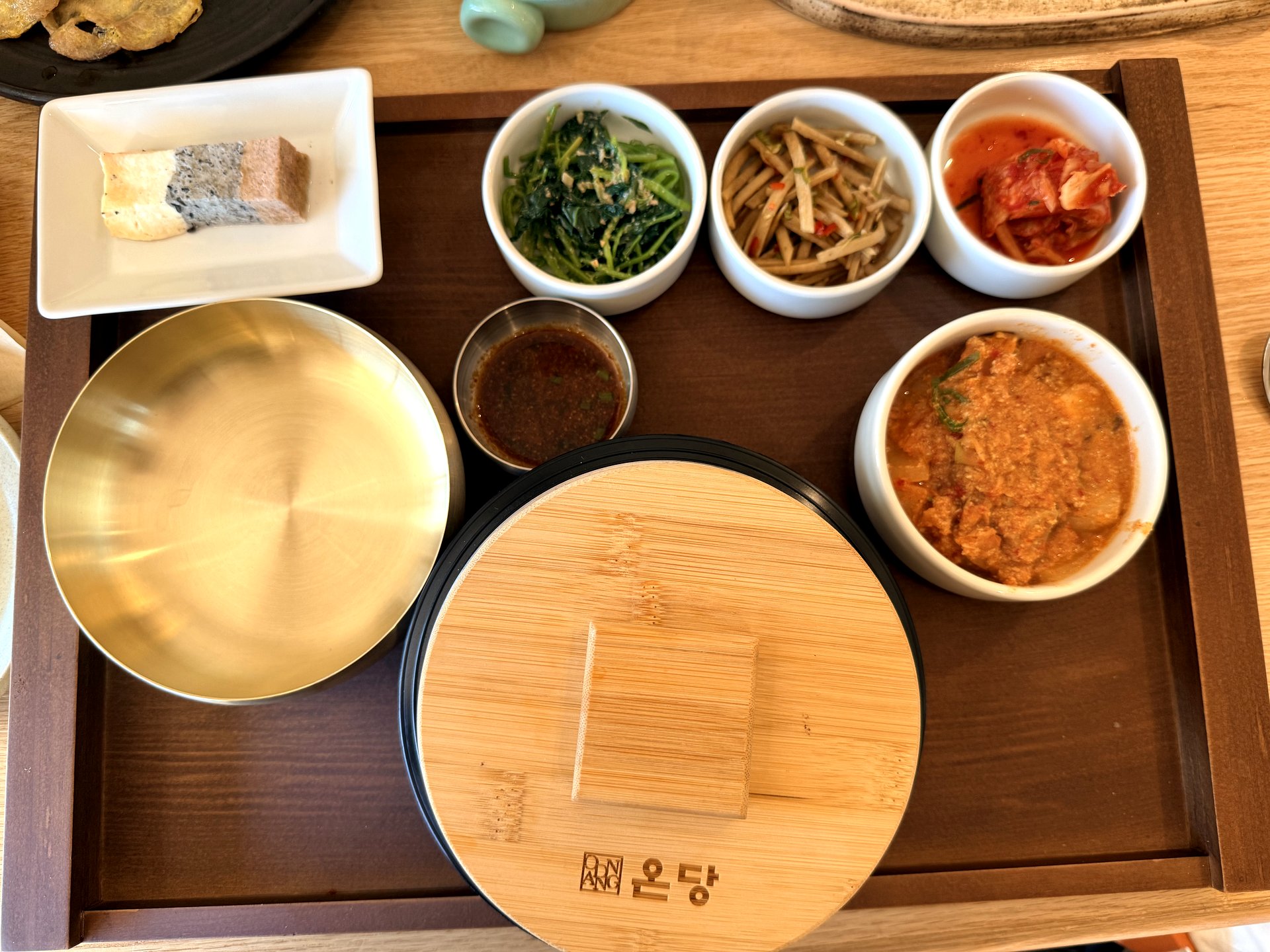 Ondang banchan tray - small white bowls of seasoned spinach, stir-fried burdock strips, kimchi and a nutty soybean-pulp side, with a bamboo-lidded pot and a small dish of sauce on a wooden tray branded ONDANG