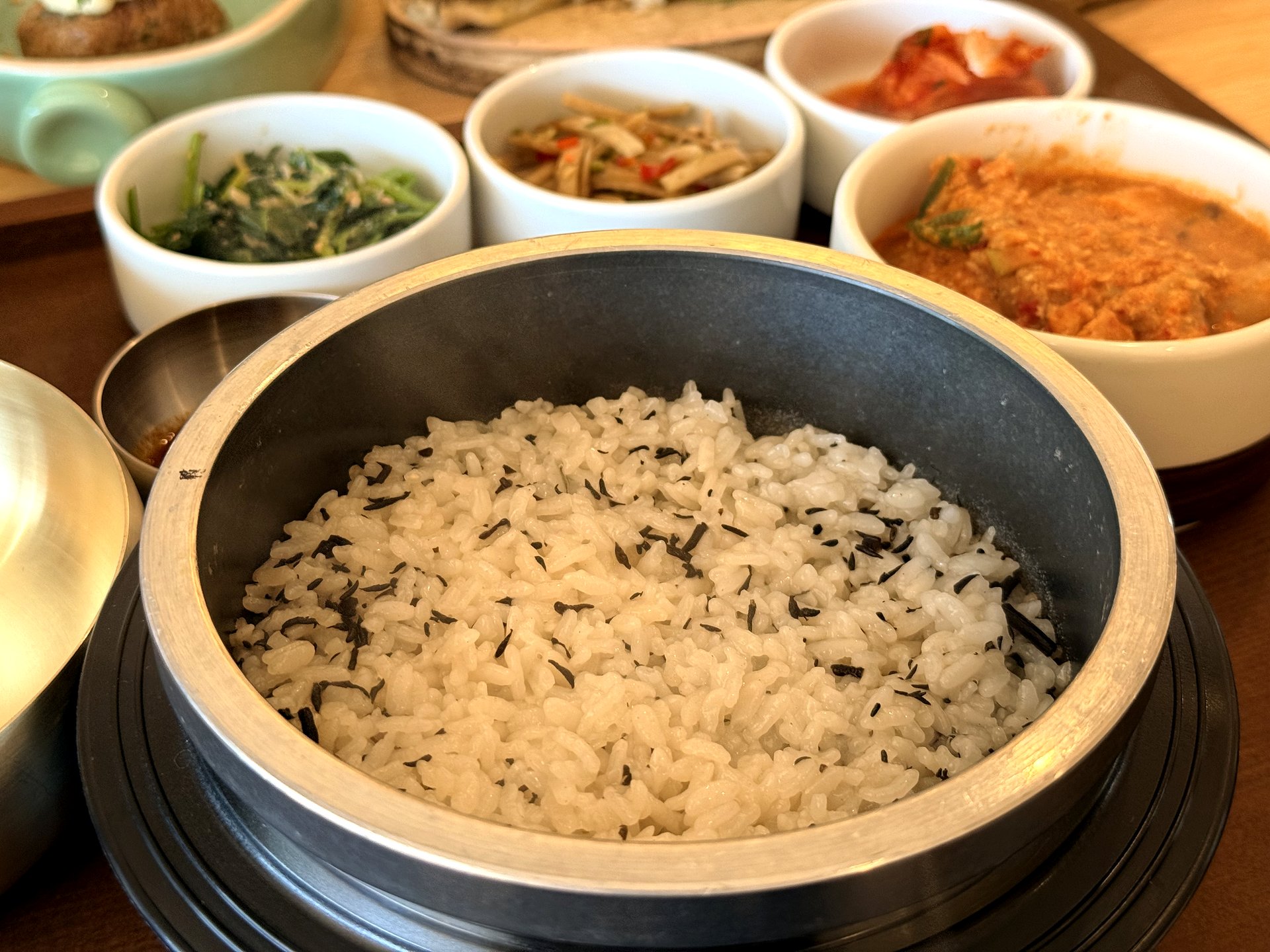 Ondang 뚝배기 밥 - freshly steamed white rice mixed with dark herb (gondeure or chwinamul) in a metal hot-pot, with banchan bowls in the background