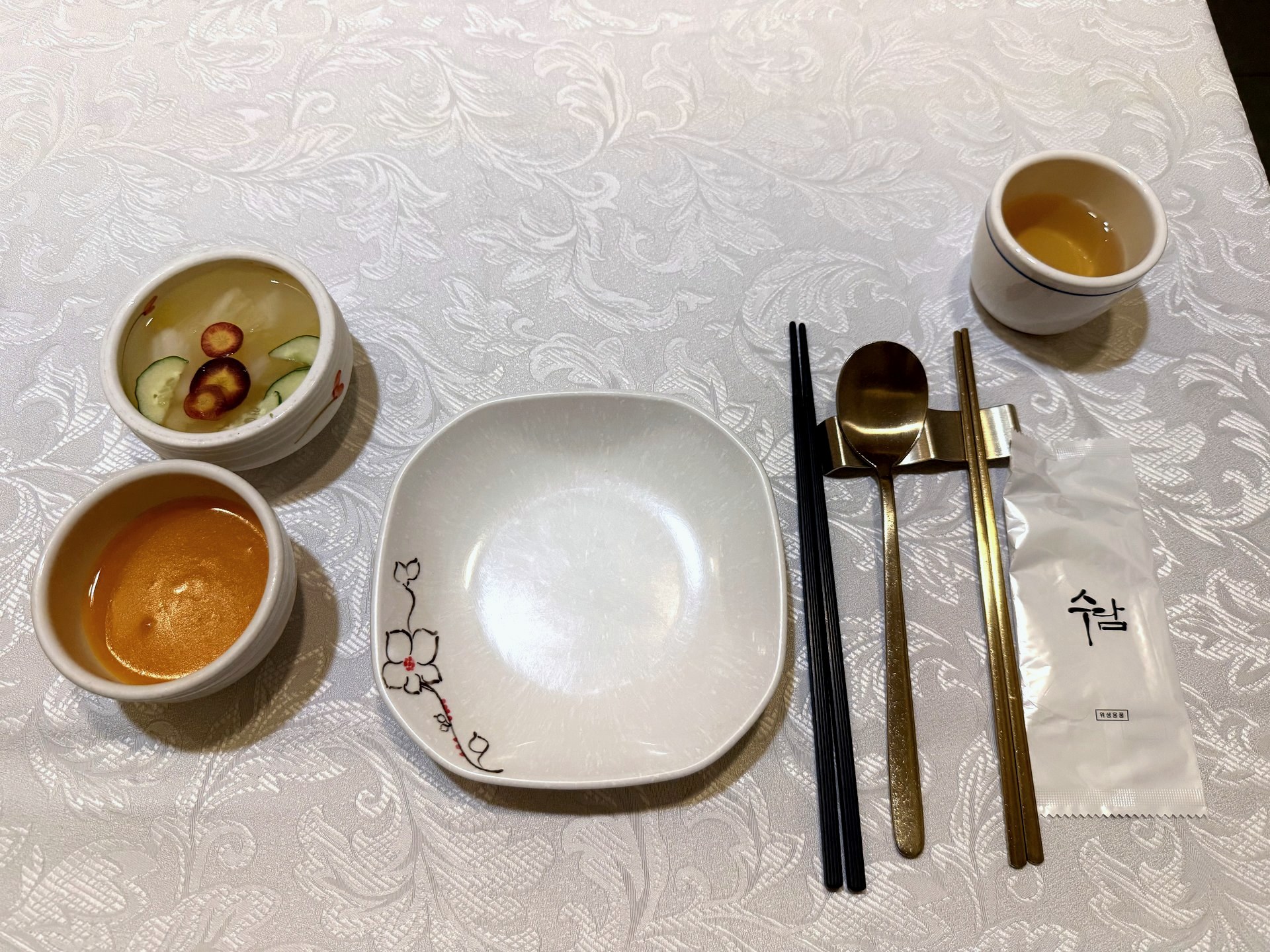 Sudam Hanjeongsik place setting - a smooth orange steamed egg custard and a light chilled appetizer in white bowls, an empty floral-rimmed plate, and brass and black chopsticks with a spoon on a rest, on a white damask tablecloth, with a Sudam-branded napkin